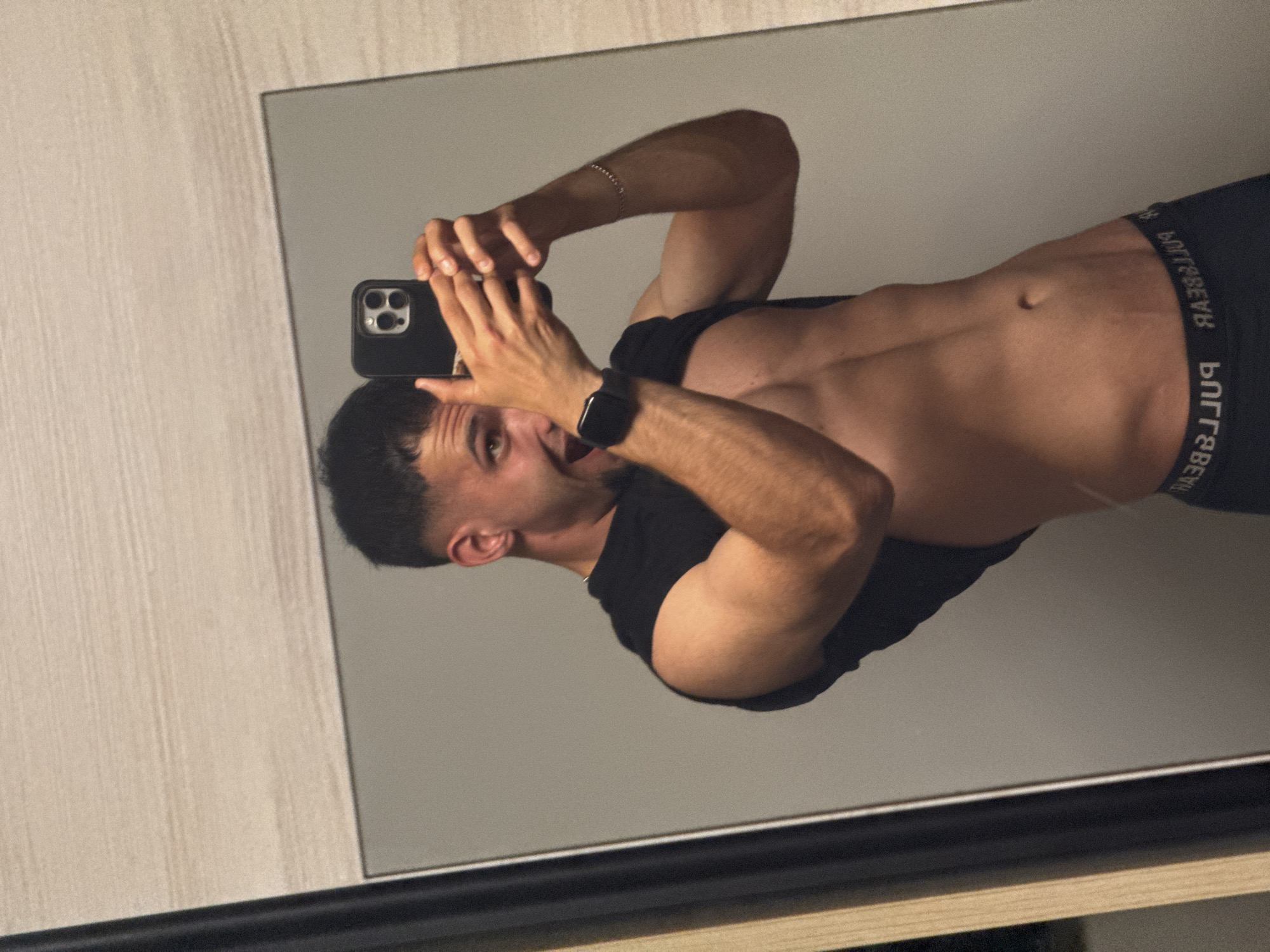 The image is a mirror selfie featuring a young, athletic man. He is positioned in front of a mirror, holding a black smartphone with a triple-lens camera towards himself to capture the reflection. He wears a black sleeveless top or t-shirt, and black boxer briefs. The brand name "PULL&BEAR" is visible in reversed text on the waistband of his boxer briefs.
The man has short, dark hair and a light stubble on his face. His mouth is slightly open, and he appears to be looking towards the camera with a relaxed expression. A silver chain bracelet is on his right wrist, and a black smart watch is on his left wrist. His torso shows visible muscle definition, particularly in the abdomen and arms.
The scene is indoors, with a light-colored, textured wall visible to the left of the mirror. The lighting is warm and generally even. No specific time of day or weather can be discerned. The provided context states the location is Shumen, Bulgaria.