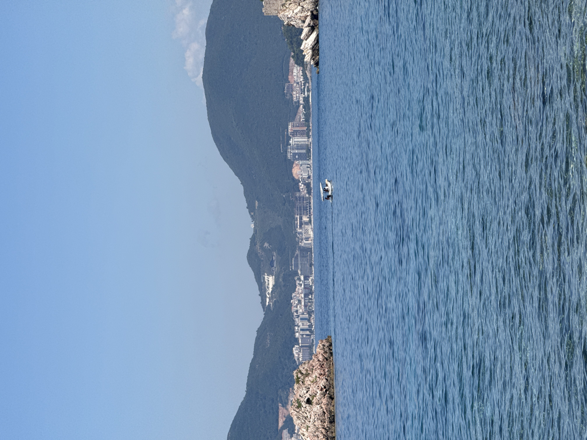 The image shows a view of the sea in front of Sveti Stefan, Montenegro. In the foreground, the blue sea has gentle ripples. In the middle ground, a small white boat with two people is visible, heading towards the right of the frame. To the left of the boat, a rocky island juts out of the water. In the background, a coastal city with numerous buildings is spread out along the shoreline, nestled at the foot of green, tree-covered mountains. A stone fortress ruin is visible on a rocky outcrop to the far right. The sky is clear and blue.