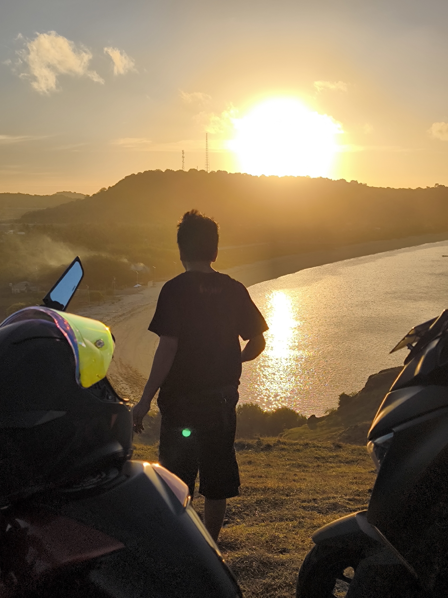 A person stands with their back to the viewer, overlooking a beach at sunset, with two motorbikes parked in the foreground. The location is Kuta, Indonesia, specifically a hillside viewpoint overlooking a beach. The person is wearing a dark t-shirt and dark shorts. A motorcycle helmet, predominantly black with a yellow visor, is visible on the left. The sun is setting over the water, creating a bright glare and illuminating the beach and the ocean. The background features a hilly landscape with trees and some visible structures, including what appears to be a telecommunications tower. The ground is covered in dry grass. A green reflective sticker is on the person's shorts.