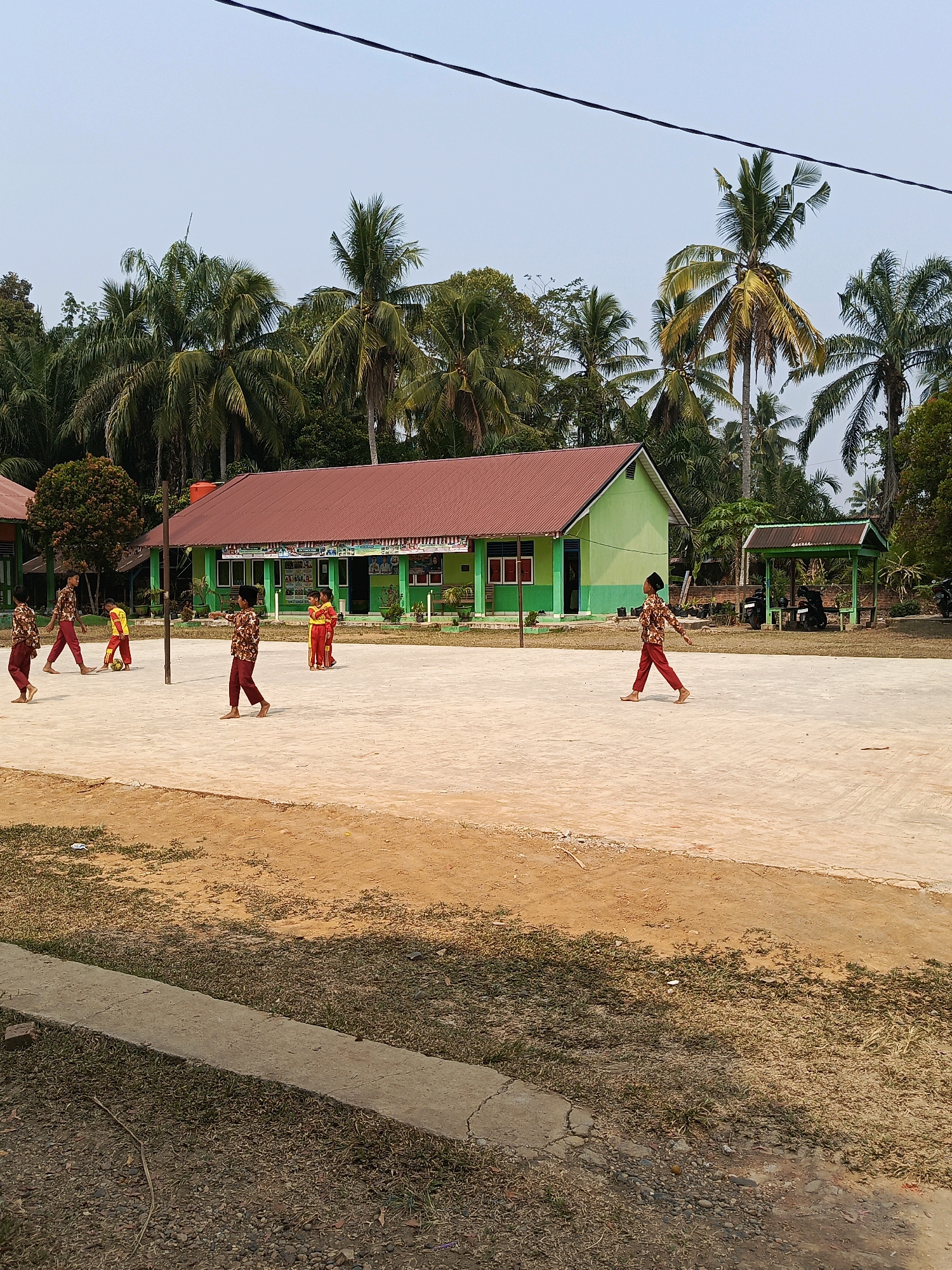 In Megang Sakti IV, Indonesia, a schoolyard scene unfolds under a clear sky, framed by lush palm trees. A green school building with a red roof dominates the background, while a smaller, open-air pavilion stands to its right, housing two parked motorcycles. The foreground is a dusty, beige-colored court, separated from a patch of dry, brown grass by a concrete curb. Several children, dressed in patterned maroon and white shirts and maroon pants, are engaged in various activities on the court. One child plays with a yellow and red ball, while others walk or stand in groups. The overall impression is of a bright, sunny day in a rural Indonesian school setting.