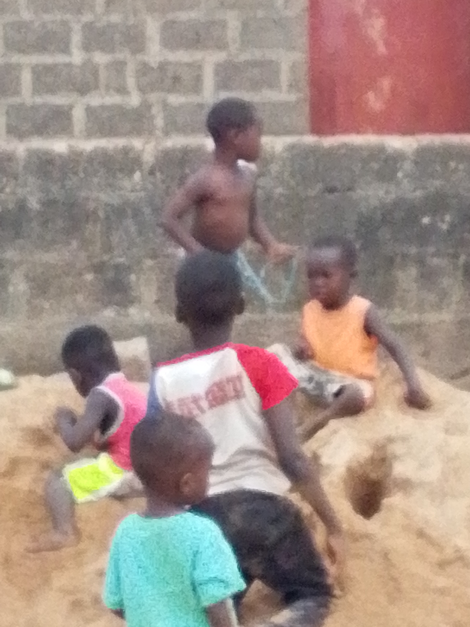 A group of five children are playing on a sandy surface in an urban environment of Unknown, Unknown. Three of the children are closer to the foreground, with the child nearest to the viewer wearing a blue shirt and facing away. Behind this child, another is seated, wearing a shirt with red and white coloring and black shorts or pants. To the left, a third child is wearing a pink top and yellow shorts. In the background, another child is sitting on the sand in an orange sleeveless shirt, and a fifth child is standing shirtless further back, near what appears to be a wall constructed of cinder blocks, with a red structure on the right.