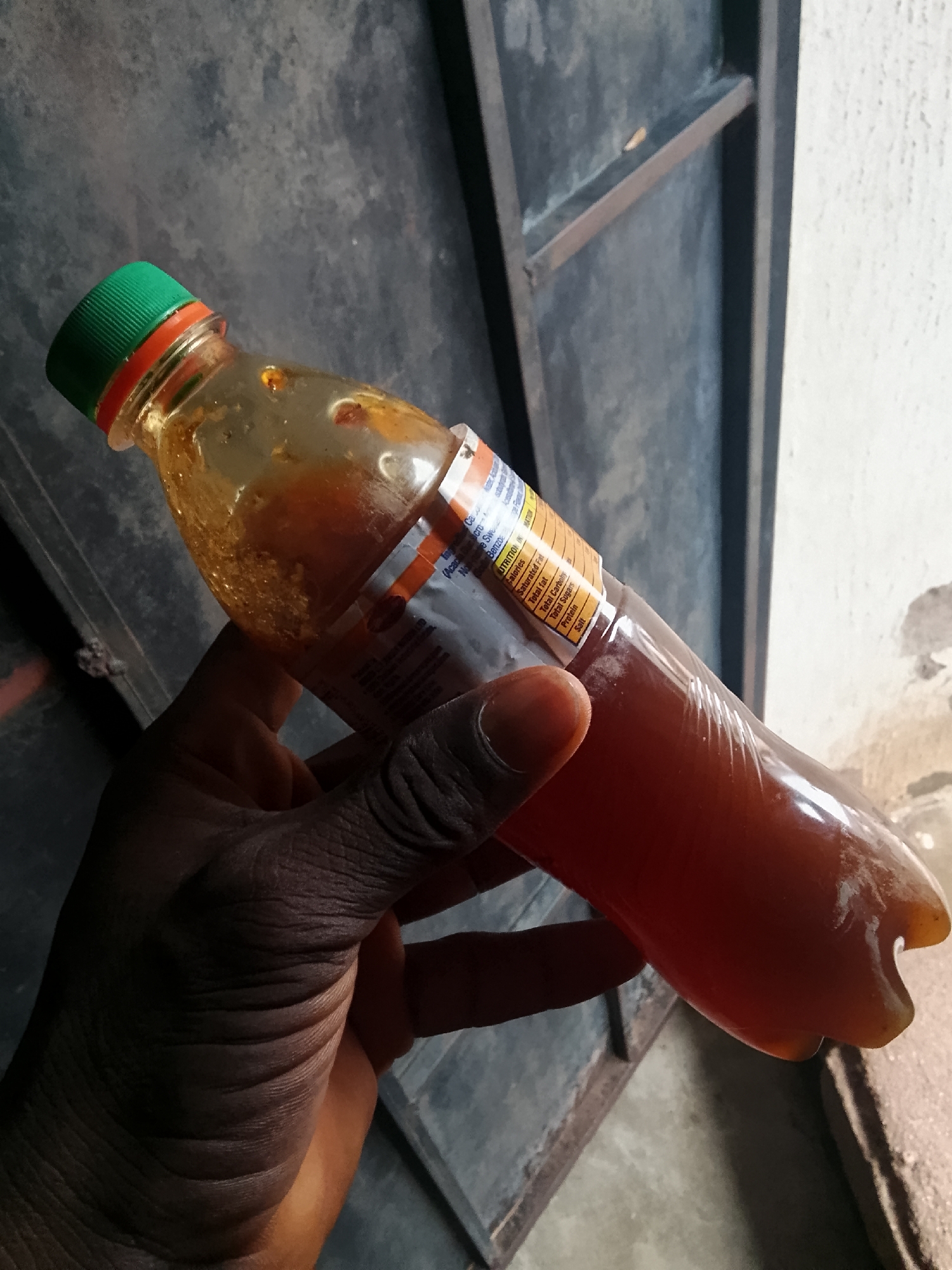 A hand with dark skin holds a partially filled plastic bottle containing an amber-colored liquid with visible particles. The bottle has a green cap and an orange and white label with nutritional information. The background consists of a dark, textured surface, possibly a wall or door, with a metal frame. The image appears to be taken in an outdoor or semi-outdoor setting.