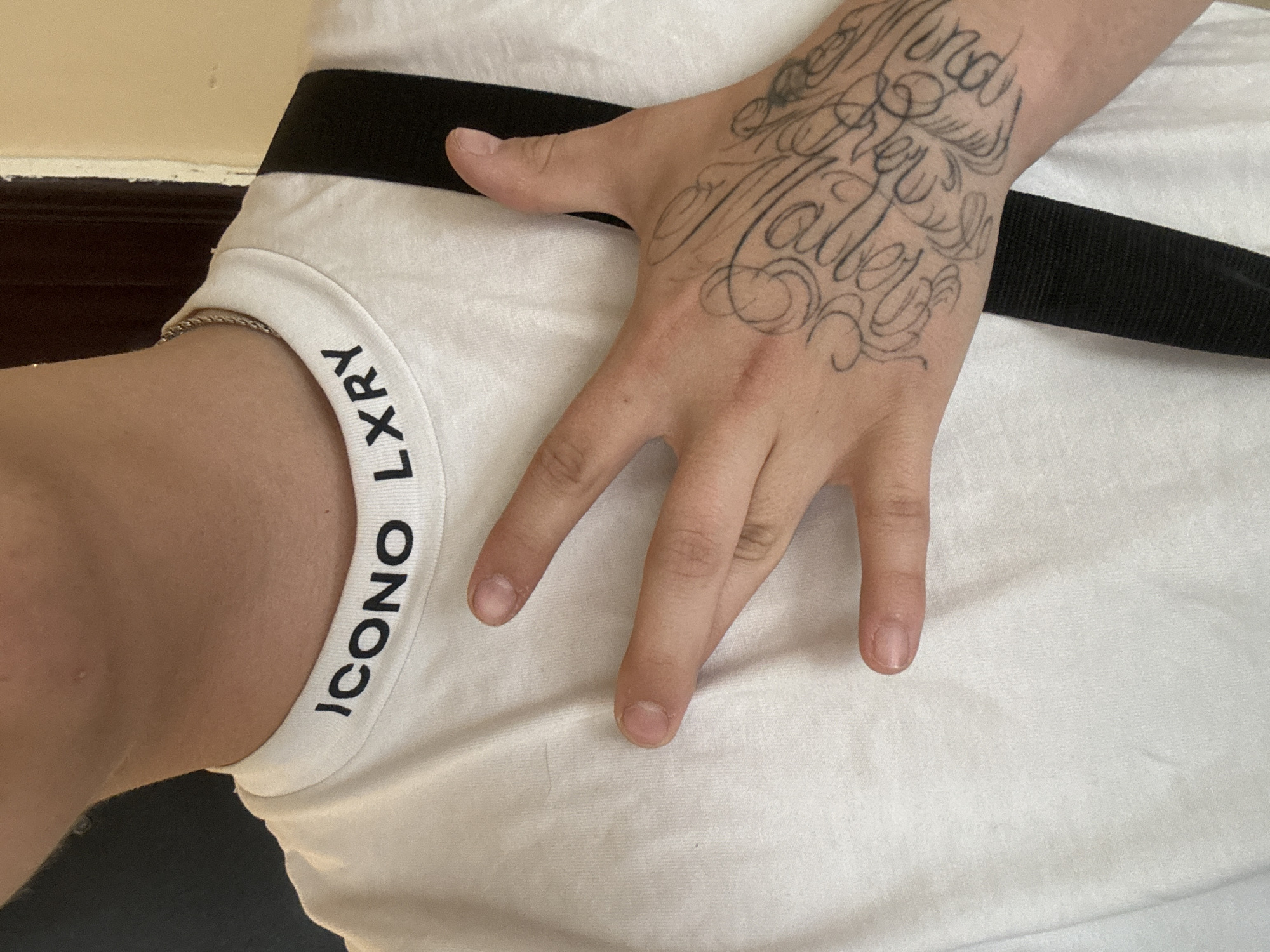 A close-up shot features a person's hand with a detailed tattoo on the back, resting on a white garment with a black stripe. The hand is lightly touching the fabric, with the fingers slightly splayed. Visible on the left side is the edge of a white undergarment with the words "ICONO LXRY" printed on it. The overall composition focuses on the texture and details of the hand, tattoo, and clothing. The location is not evident from the image.