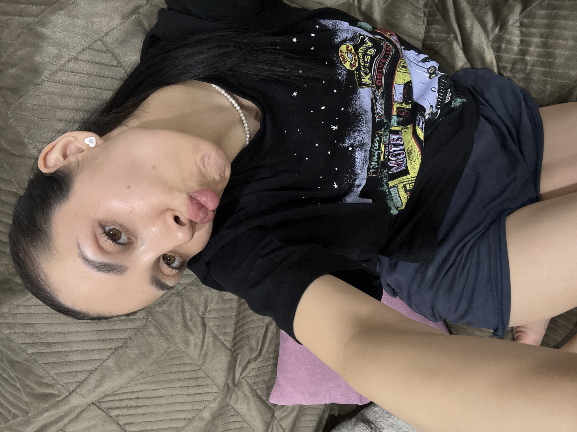 A person is lying on a textured brownish-grey blanket or bedspread in what appears to be an indoor setting. The individual is positioned diagonally, with their head tilted to the left. They have dark hair, light skin, and brown eyes, looking directly at the camera with a playful expression, featuring puckered lips often described as a "fish face."
The person is wearing a black t-shirt with a colorful graphic print on the front. The print depicts retro-style signs and imagery, including visible text such as "Kreamy Krisp," "DRIVE-IN," and "MOTEL." The t-shirt also features white speckles or stars. A delicate pearl necklace is worn around the neck, and a small, heart-shaped stud earring is visible in the right ear. Dark shorts are worn on the lower body.
A small pink cushion or pillow is visible beneath the person's left arm/side. In the lower right corner, parts of the person's feet or toes are faintly visible. The overall mood is casual and relaxed, suggesting a candid or selfie-style photograph. The image was taken in Votkinsk, Russia.