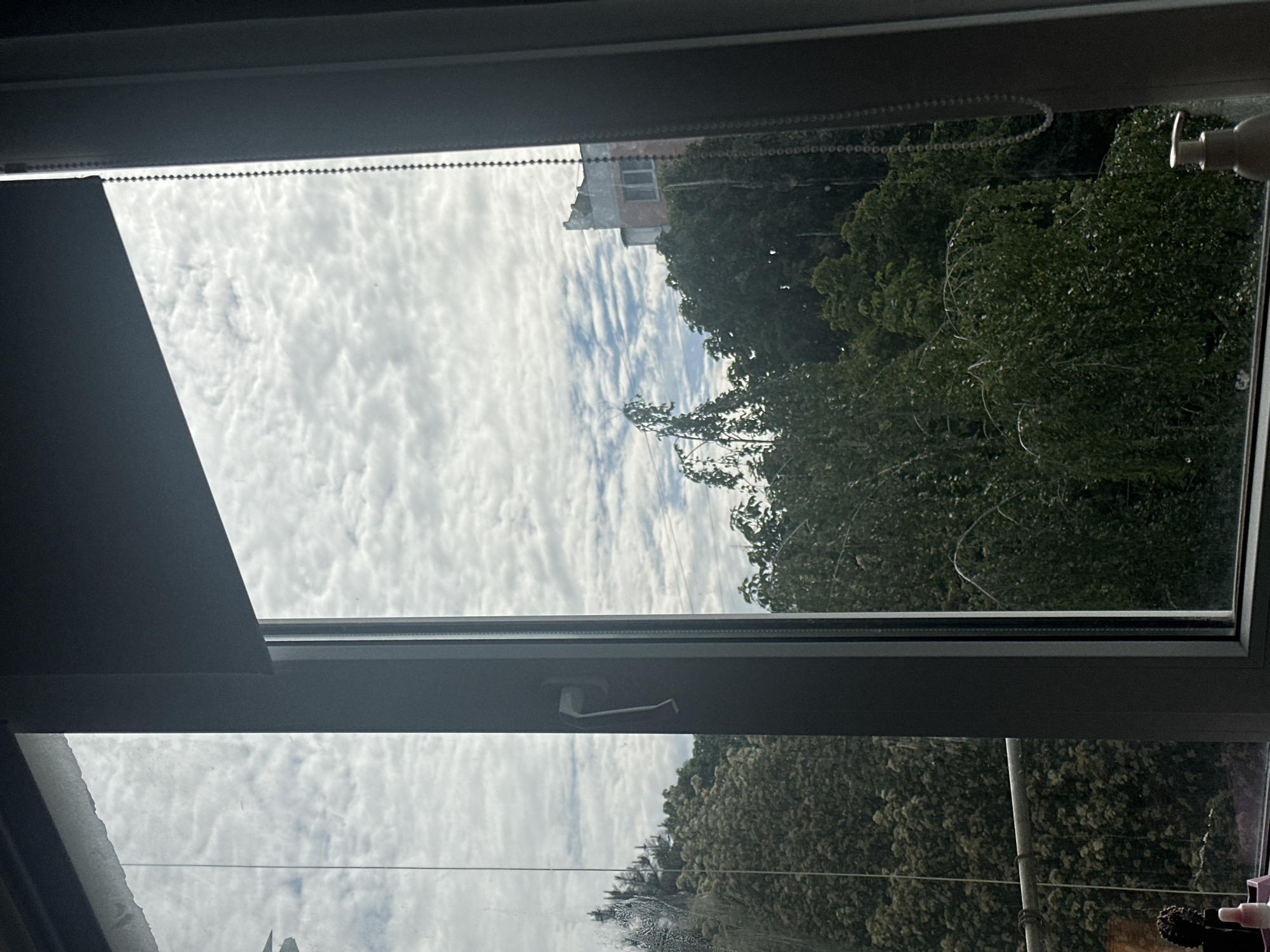 The media file displays a view looking out of a window, presented in a sideways orientation. If rotated 90 degrees counter-clockwise, the image would show a natural landscape view with the sky above and ground elements below.
In the foreground, a modern grey window frame, likely made of PVC, borders the view. A white window handle is visible on the central horizontal bar of the frame. From what would be the top-left corner of the window (in natural orientation), a dark grey roller blind is partially drawn down, with its characteristic beaded control chain running along the top edge of the window frame.
The exterior scene features a predominantly bright sky, filled with textured, patchy white clouds, resembling altocumulus or stratocumulus formations, indicating an overcast but well-lit daytime. Below the sky, a dense canopy of lush green trees fills a significant portion of the view, suggesting a summer or late spring season. Partially obscured by the foliage, a multi-story brick building with visible windows can be seen in the distance, appearing somewhat aged.
There are no people or specific activities taking place. No discernible text is visible within the image. The scene conveys a calm, natural setting consistent with the provided location context of Stavropol, Russia.