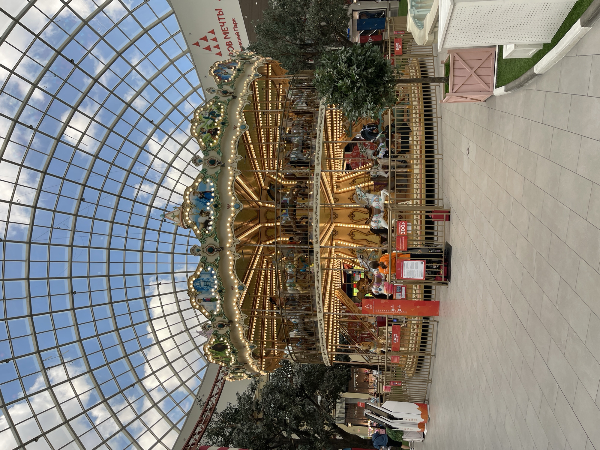 The image shows a well-lit carousel inside a large indoor space with a glass ceiling, suggesting a shopping mall or entertainment center. The carousel is ornately decorated with lights and has multiple tiers with various carousel animals, such as horses. People are visible riding the carousel and some are standing around it. In the background, there are trees, shops, and signage, including a sign with Russian text that appears to indicate an exit. The overall environment is clean and modern, with tiled floors and a light-colored ceiling structure. The presence of an indoor carousel and retail spaces suggests an urban, commercial setting.
