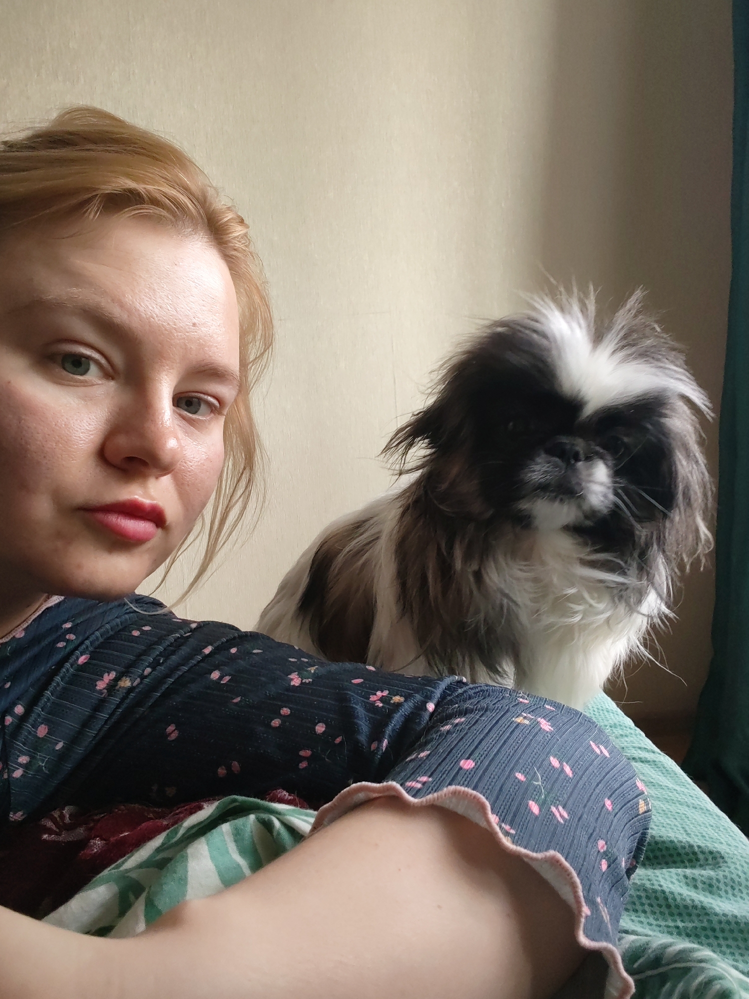 A woman is shown in a selfie-style image indoors with a dog. The woman has fair skin, blonde hair pulled back, and is wearing a dark blue shirt with pink floral patterns and a ruffled neckline. She has neutral facial expression.
Next to the woman is a small dog with long, black and white fur. The dog is gazing forward.
The background consists of a beige wall and a green curtain. The room is lit with soft, diffused light.
The image does not provide any information that could confirm the location to be Izhevsk, Russia.