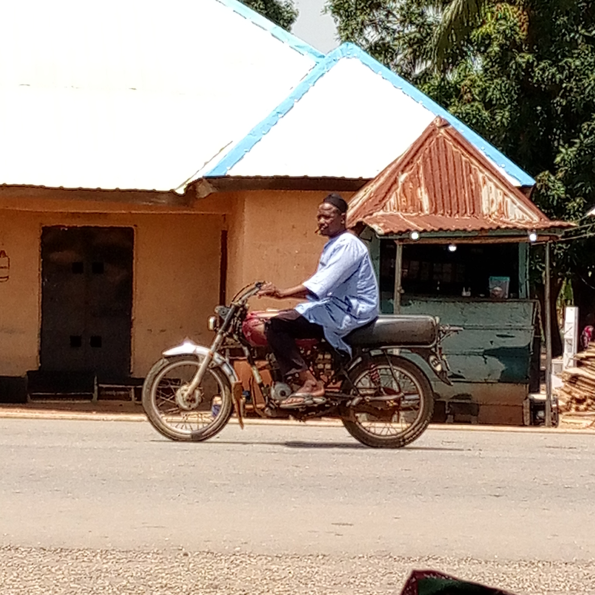 A man is riding a motorcycle on a paved road in Mararaba, Nigeria. He is wearing a light blue robe and black pants, and he has a black cap on his head. He is also wearing sandals. The motorcycle is red and black, and it has a white front fender. The man is riding the motorcycle in front of a small building with a white and blue roof. Next to the building is a rusted shed-like structure. There are trees in the background.