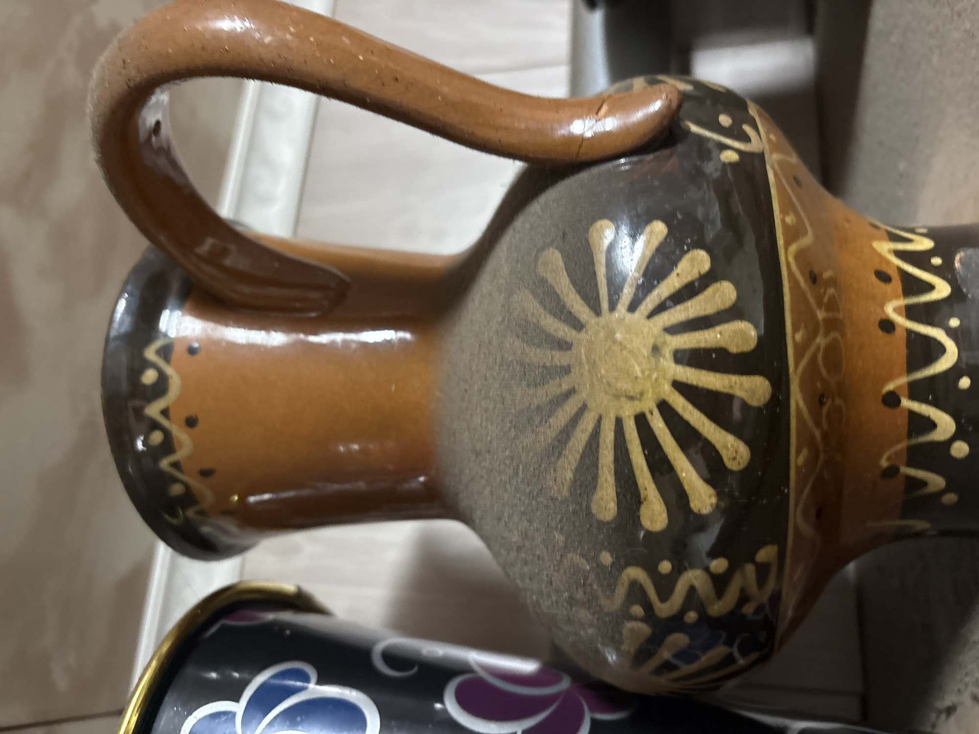 An ornamental, dusty, brown and black ceramic jug with a handle is the central focus of the image. The jug has yellow, sun-like and wavy patterns. To the left of the jug, a portion of a black vase with blue and purple floral designs is visible. The objects are positioned on a shelf or counter, with a light-colored wall or background behind them. The overall impression is of decorative items that are likely in storage or display, showing signs of dust accumulation. The image was taken at a domestic location in Ветлужский, Russia.