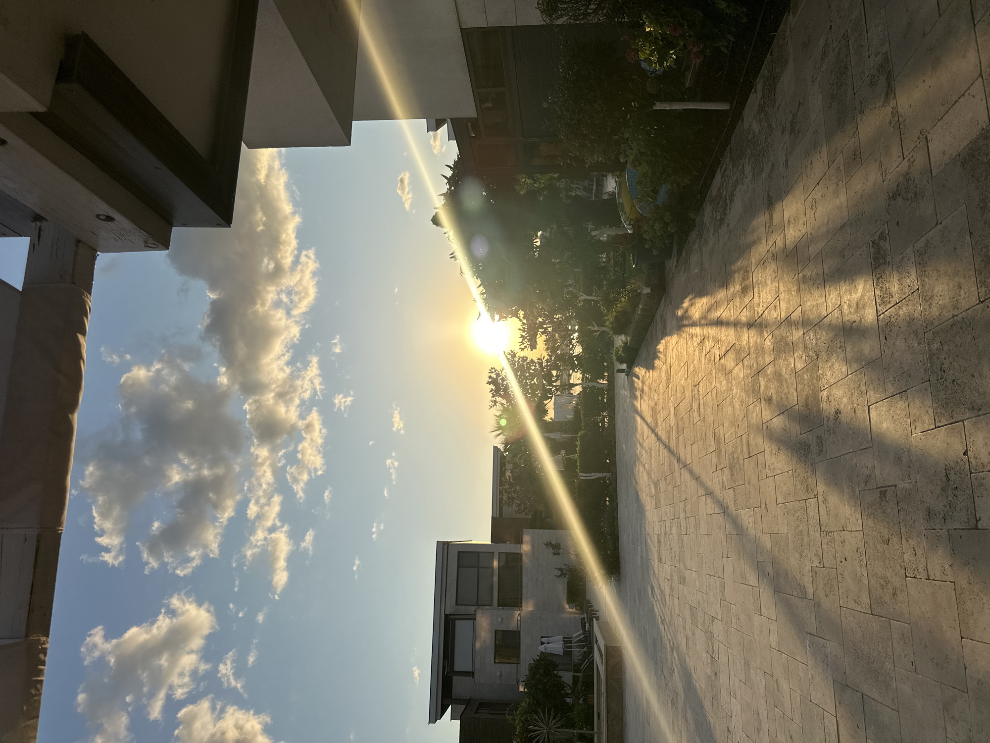 The image captures a sunset over a modern residential area in Sozopol, Bulgaria. The scene is characterized by a paved courtyard with large, rectangular stone tiles. Long shadows are cast across the courtyard by unseen objects, extending from the right side of the frame towards the left.
On the left, a two-story modern building with large windows and a flat roof is visible. In front of it, there are lounge chairs and closed umbrellas, suggesting an outdoor seating area. Lush greenery and trees are interspersed throughout the scene, with a distinct line of dense bushes bordering the courtyard on the right.
The sun is low in the sky, positioned towards the center-left of the image, and is creating a bright lens flare that streaks diagonally across the frame. The sky is a clear blue with scattered, fluffy white clouds illuminated by the setting sun. The overall atmosphere is serene and warm, typical of a late afternoon or early evening in a coastal town. There are no people visible in the frame.