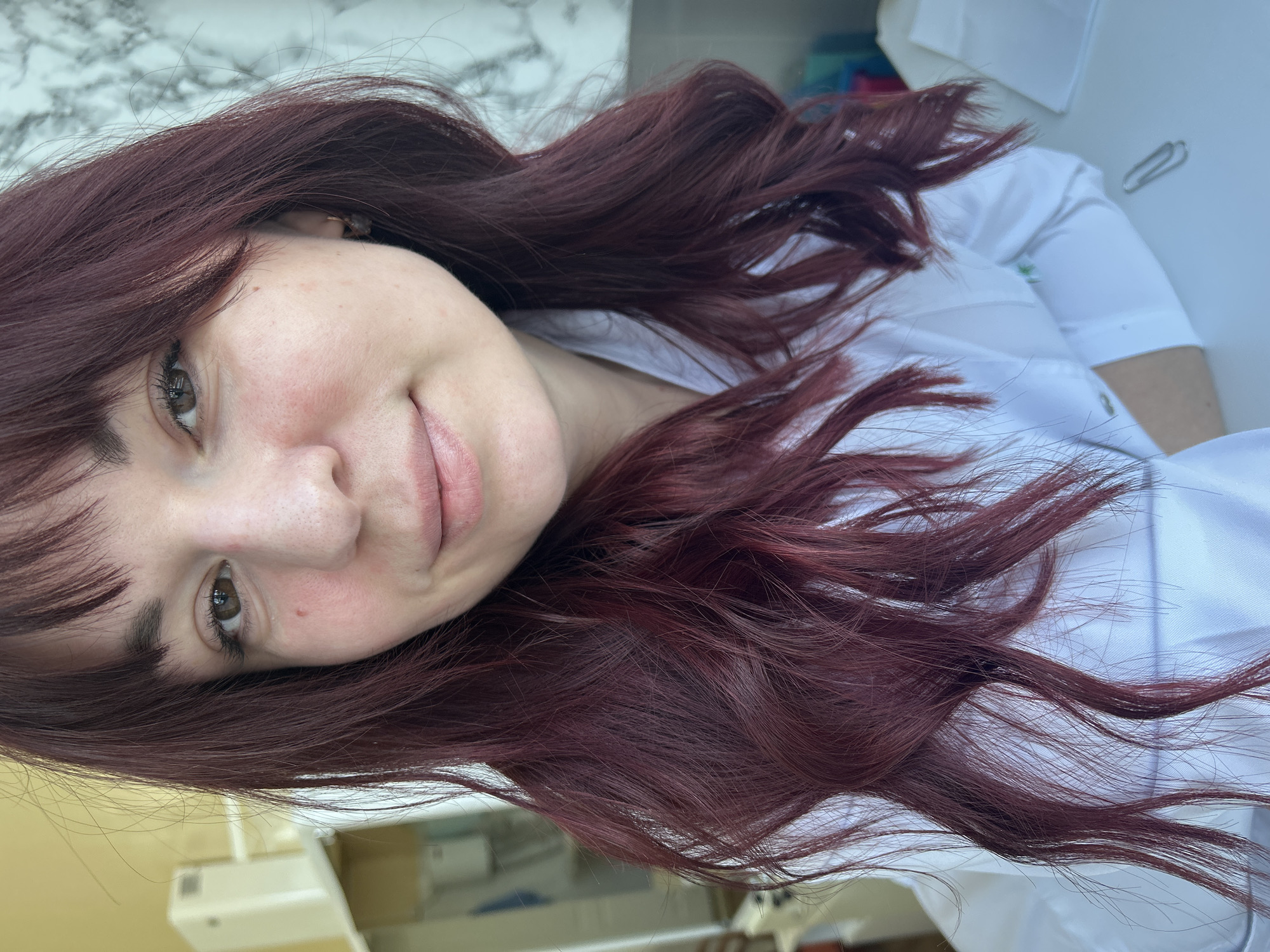 A woman with long, wavy, reddish-brown hair, a fair complexion, and light brown eyes is pictured indoors. She is wearing a white shirt. A paperclip is visible on the table to the right. The background includes a surface with a marble-like pattern and hints of other objects. There is no evidence in the image to associate it with Фоки, Russia.