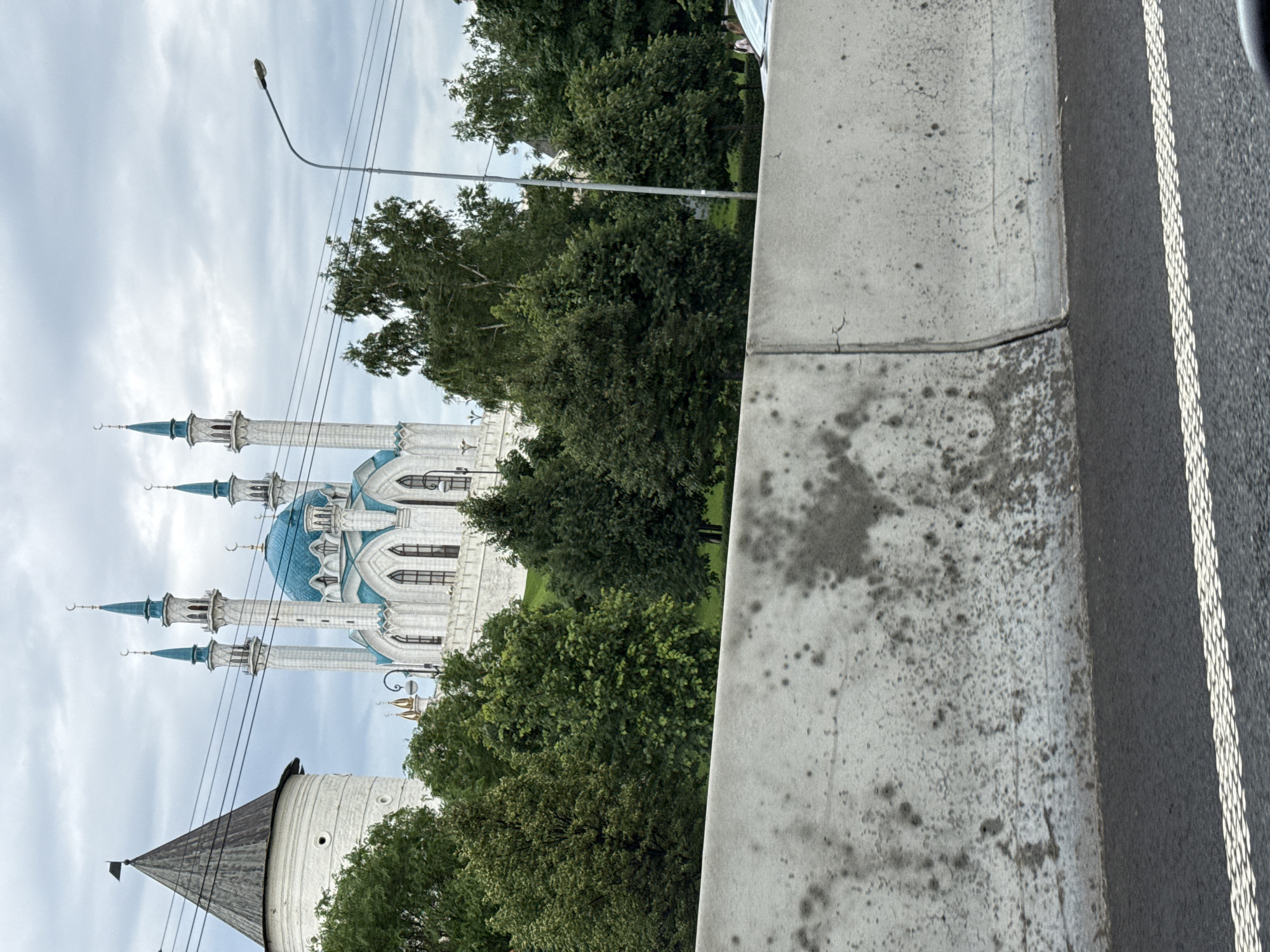 The image shows a view of the Kul Sharif Mosque in Kazan, Russia. The mosque, with its white walls, blue domes, and tall minarets, is partially visible behind a dense cluster of green trees. The sky above is overcast with grey clouds, suggesting a cloudy day. To the right of the mosque and trees, a portion of a paved road with a white dashed line is visible, indicating an urban or roadside setting. A concrete structure, possibly a curb or barrier, runs along the edge of the road. The image is taken from a low angle, with the camera tilted upwards. The overall impression is of an architectural landmark set against a natural and urban backdrop on a seemingly quiet day.