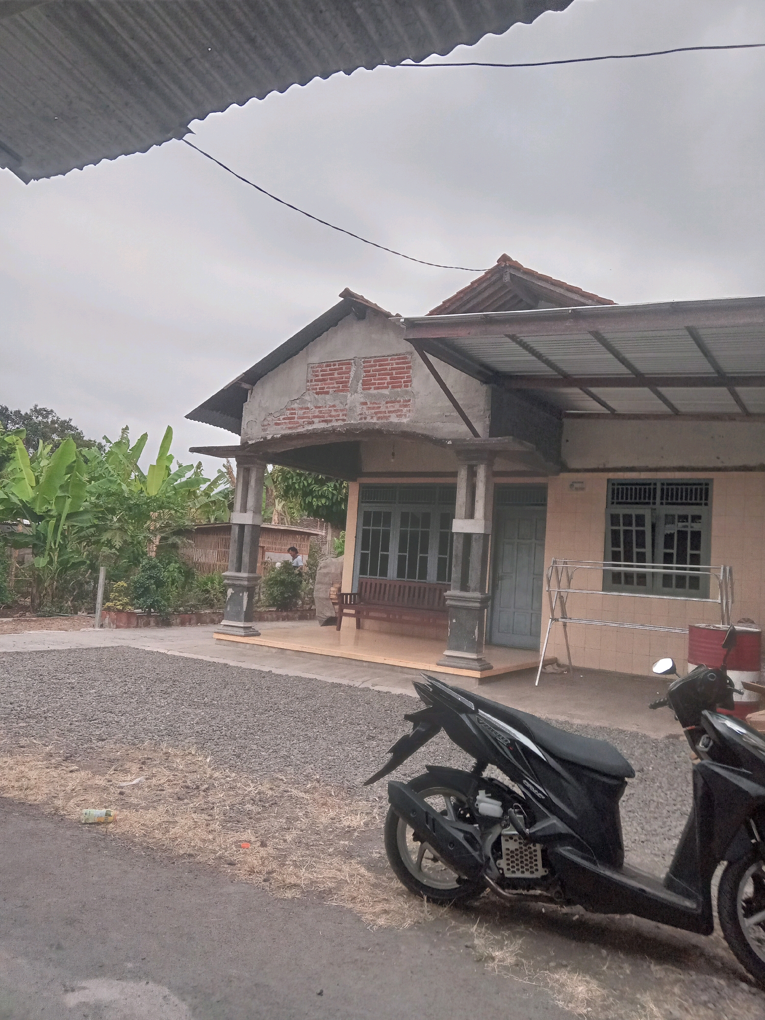 The image depicts a street scene in DESA KLAHANG, Indonesia. In the foreground, a black motorcycle is parked on the side of an asphalt road. The road is bordered by dry grass and gravel. Behind the motorcycle, a gravel driveway leads to a single-story house. The house has a light-colored tiled facade and features large windows with a grid pattern. A covered porch extends from the front of the house, supported by pillars. To the left of the house, a lush garden area is visible, with numerous banana trees and other vegetation. In the background, a cloudy sky is present. A person can be seen standing near the garden area. The overall scene suggests a residential area in a rural or semi-rural setting.