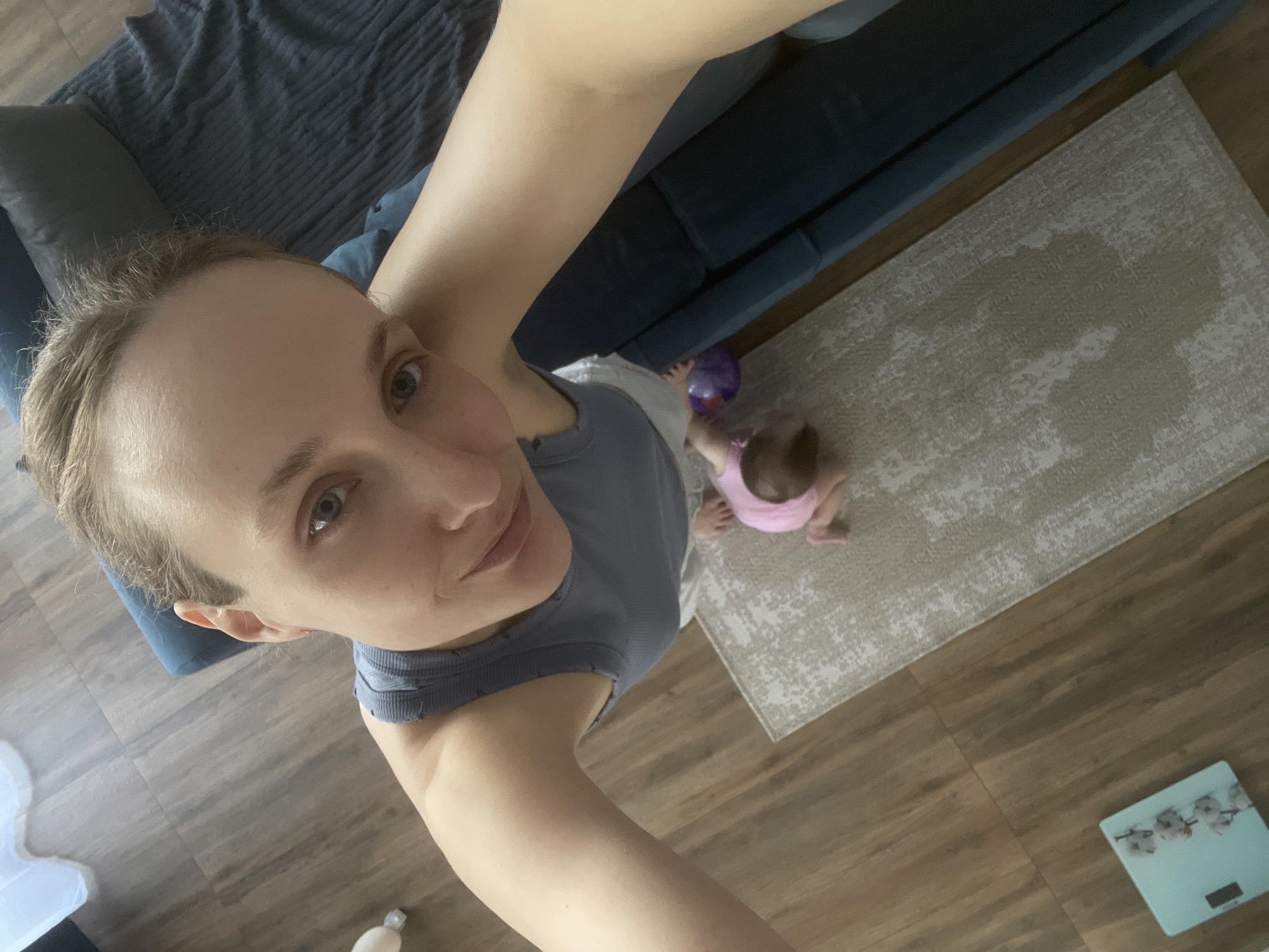 A high-angle selfie taken indoors. A fair-skinned woman with short hair is the primary subject, looking at the camera. A baby, wearing a pink outfit, is on a rug. A blue couch is partially visible. A purple ball lies near the baby. A scale is on the wooden floor. The location is possibly in Подшивалово, Russia.