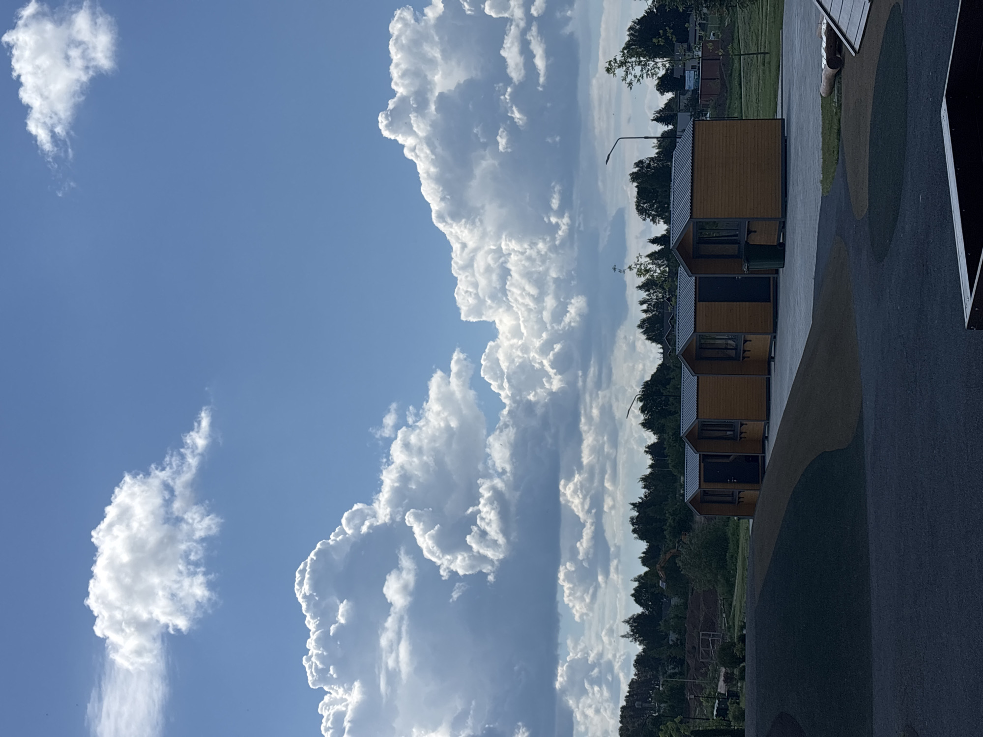 The image presents a daytime scene in Varaksino, Russia, under a bright blue sky populated with prominent, billowing cumulus clouds. In the foreground, aligned horizontally along the right edge, are several identical wooden cabins with dark grey roofs. The cabins are situated on a paved area with colorful circular patterns. Beyond the cabins and pavement is a line of trees with dense green foliage. In the distance, partially obscured by trees, are glimpses of other structures and greenery. The overall impression is that the cabins are located in a recreational or park area.