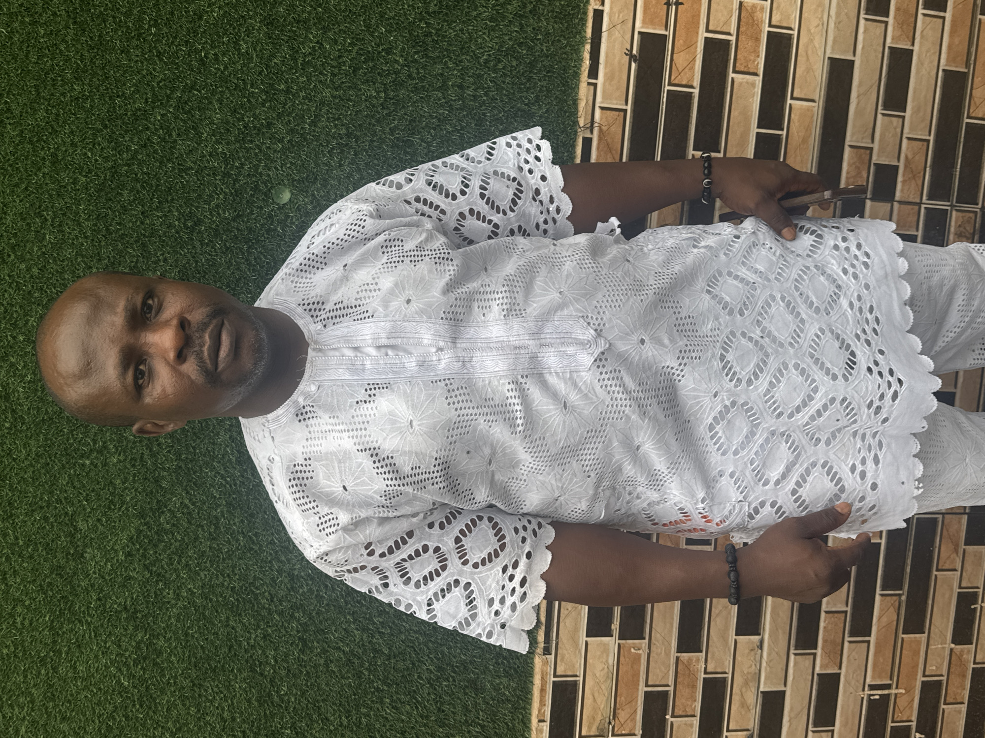 The image shows a man wearing a white, patterned, short-sleeved top and matching pants. He is positioned between two surfaces: a patch of green artificial turf on his left and a wall covered in brown and beige rectangular tiles on his right. He is wearing a black beaded bracelet on each wrist. The image appears to be taken in Nkpor, Nigeria, as indicated in the prompt.