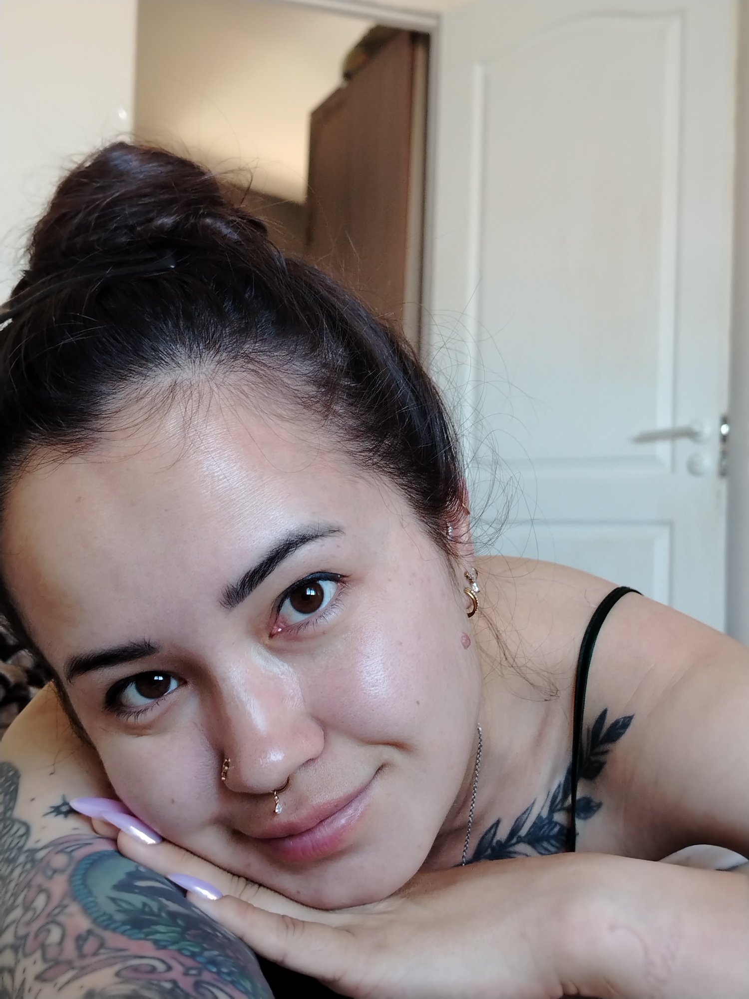 The image shows a close-up, chest-up portrait of a young woman with dark hair pulled up in a bun. She is lying down, resting her chin on her hands. She has several piercings, including a septum ring and an earring. She also has tattoos on her arms and chest, including a floral design on her chest and a large, colorful tattoo on her left arm. The background appears to be a bedroom, with a white door and a wardrobe visible. The lighting is natural, suggesting it might be daytime. The exact location in Noviy, Russia, cannot be determined from the visual information provided.