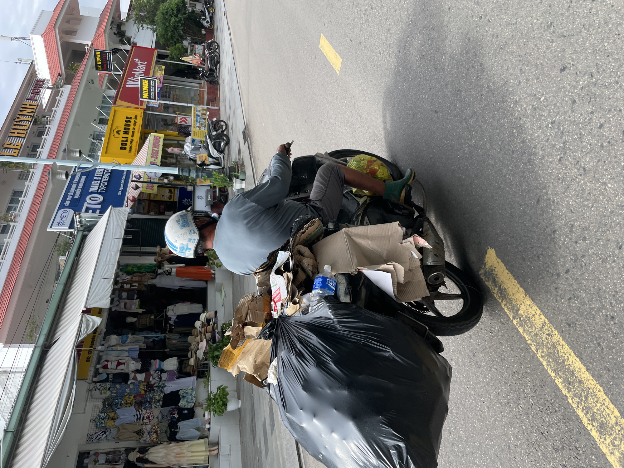 A man on a heavily loaded scooter occupies the mid-ground of a street scene in Phu Thuy Ward, Vietnam. He is dressed in a light blue long-sleeved shirt, dark shorts, green boots, and a white helmet, and appears to be holding a small object in his right hand.
The scooter is laden with a substantial collection of items, including a large black trash bag, numerous cardboard boxes, and various brown paper bags, suggesting a role in recycling, waste collection, or deliveries. A blue "POCARI SWEAT" bottle is visible atop the piled goods.
The foreground consists of an asphalt road with a distinct yellow painted line. In the background, a row of commercial buildings lines the street, featuring awnings and displays of clothing items. Various Vietnamese business signs are prominent, such as "LÊ HUỲNH", "WinMart+", and "DOLI HOUSE" (which advertises "TRANG PHỤC - MAKE UP - CHỤP ẢNH" or "Clothing - Makeup - Photography"). Another sign for "METO TRAVEL & EVEN ТУРОПЕРАТОР" (Tour Operator) displays an address "135A Nguyen Dinh Chie M". Several other motorbikes are parked along the sidewalk in front of these establishments.
The bright daylight indicates a daytime setting, capturing a typical bustling commercial street in Vietnam, characterized by local businesses and motorbike activity.
