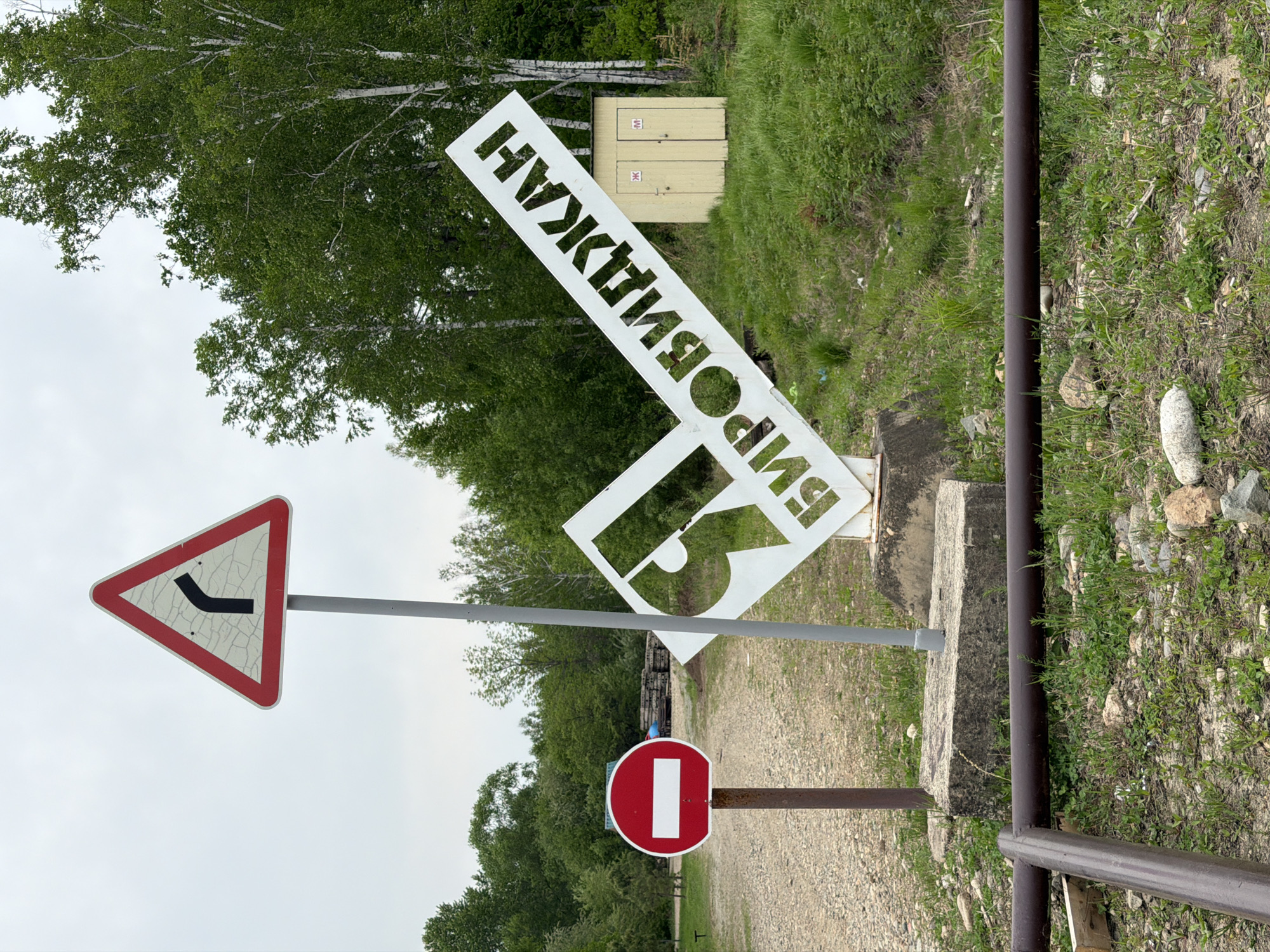 The image captures a scene in Birobidzhan, Russia, featuring a collection of roadside signs and a general outdoor environment. The signs include a triangular road sign with a left turn indicator, a circular red and white 'no entry' sign, and a large, white, Cyrillic sign that spells out "Birobidzhan". The environment appears to be alongside a road or path, with grass, trees, and a small utility box in the background. A metal railing is visible in the foreground. No people are visible.