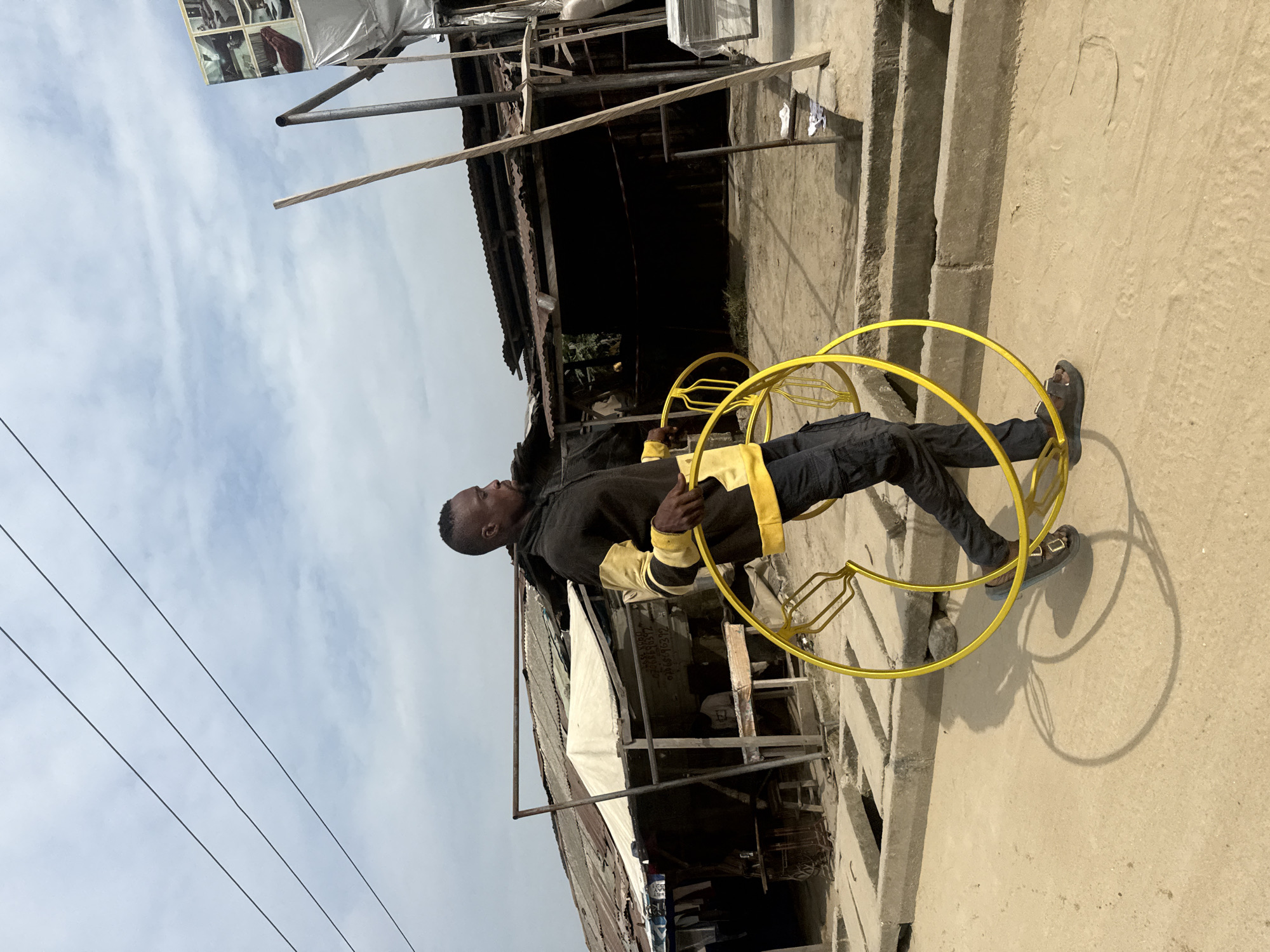 The media file presents a young Black man standing outdoors in a bright, informal urban setting in Ado, Nigeria. He wears a black long-sleeved top with yellow accents, dark trousers, and dark sandals. In each hand, he holds a large, bright yellow, circular metal frame that extends to the ground, casting distinct shadows on the sandy concrete surface, indicating strong daylight. The man is positioned slightly sideways, looking towards the left, appearing focused on the apparatus he holds.
The background reveals a bustling, somewhat makeshift commercial environment. Behind the man is a structure featuring corrugated metal roofing, wooden beams, and concrete elements, possibly steps or walls. A wooden sign is visible on one of these structures, displaying the text "ZIDAN AUTO SPARE" along with phone numbers "08080088587" and "07038556332", identifying it as an auto parts or repair shop. The sky above is light blue with scattered white clouds, and thin power lines are visible in the upper left corner. A subtle detail on the ground near his feet includes a faint heart-like shape drawn in the dust. The man's expression is neutral, and the overall scene captures an ordinary moment in this vibrant local setting.