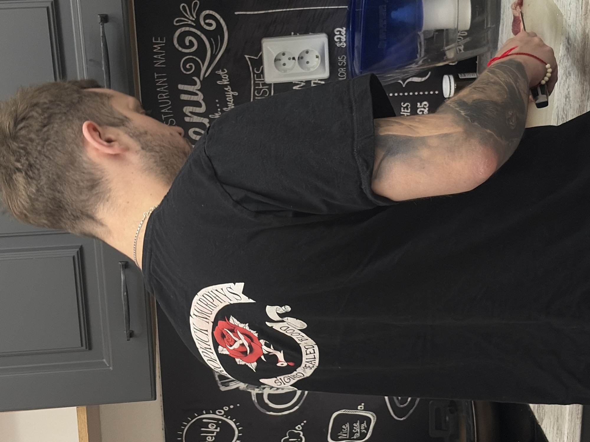 A man with short brown hair and a beard is captured from behind and slightly to the side, facing away from the viewer. He wears a black short-sleeved t-shirt featuring a distinctive white logo on the back: "DROPKICK MURPHYS" arched above a red rose, with "SIGNED SEALED BLOOD" below. A silver chain necklace is visible around his neck.
His right arm, heavily tattooed with dark, possibly blackwork or shaded designs, is extended. He wears a red string bracelet with white beads on his right wrist and is holding a dark-handled knife, appearing to cut a piece of light-colored food or material on a white surface. This activity suggests he is engaged in food preparation, likely in a kitchen.
The background features dark grey kitchen cabinets with black bar handles on the left. To the right, a blackboard-style wall is visible, adorned with chalk drawings and text. Notable text includes "RESTAURANT NAME," "menu," phrases like "h... ways hot," "ISHES," "hello!," and "Nice to see you." Some pricing, like "$22" and "25," is also present. A white European-style electrical outlet with two circular sockets is mounted on the blackboard. Behind the man's arm, a blue and white plastic container (possibly a water filter or pitcher) and a partially visible dark bottle are seen. The presence of the European electrical outlet aligns with the Saint Petersburg, Russia context. The scene suggests a casual setting, possibly a home kitchen or a small eatery.