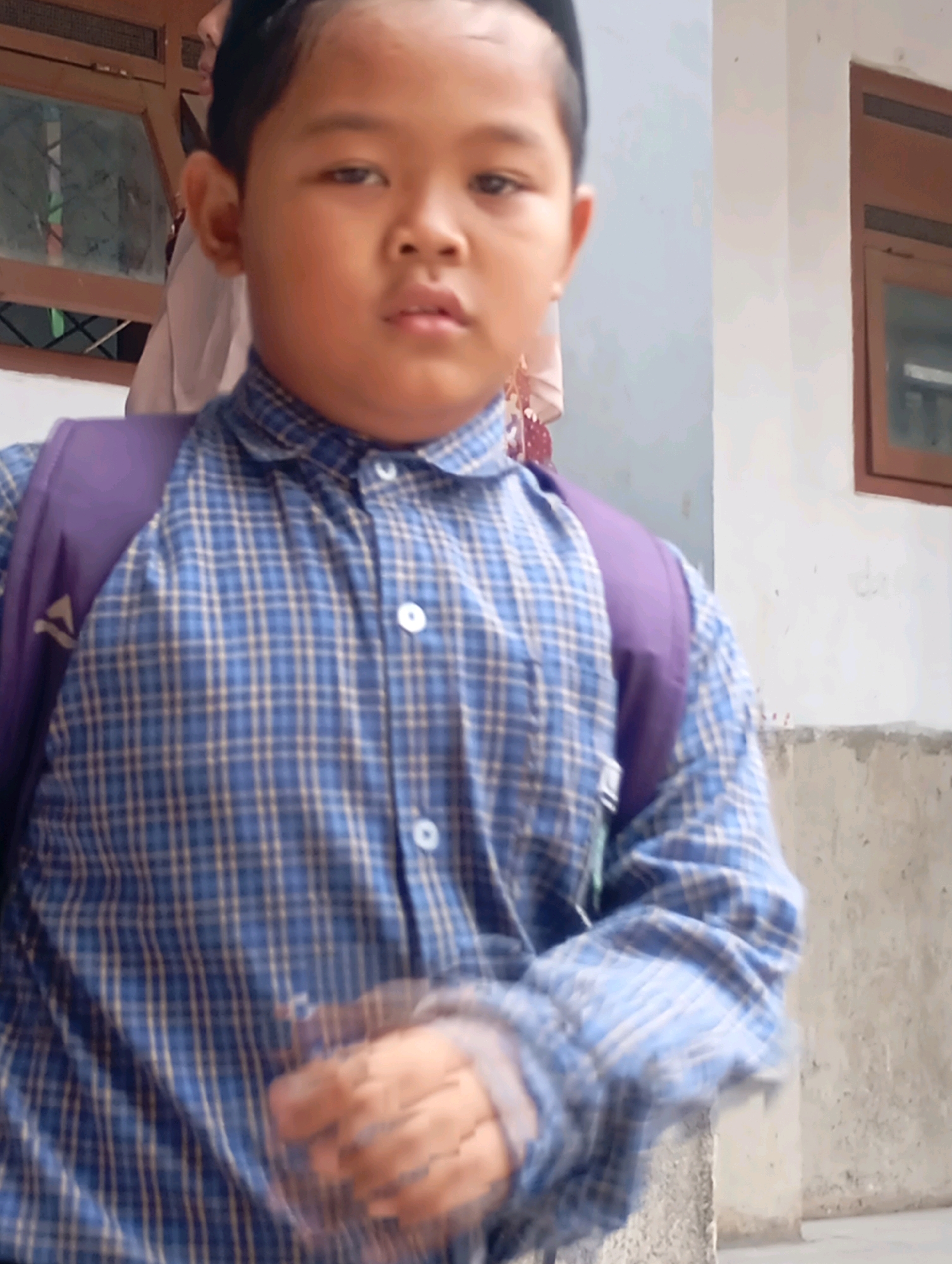 The image shows a young person wearing a blue and white plaid collared shirt and a purple backpack. The person is facing forward and looking at the camera. The background appears to be outdoors, with a building that has a wooden-framed window and a concrete wall. The location is identified as Babelan, Indonesia. The person's left hand is blurred due to motion. Another person, partially obscured, is visible behind the main subject, wearing a light-colored headscarf. The scene appears to be during the daytime, with natural lighting.