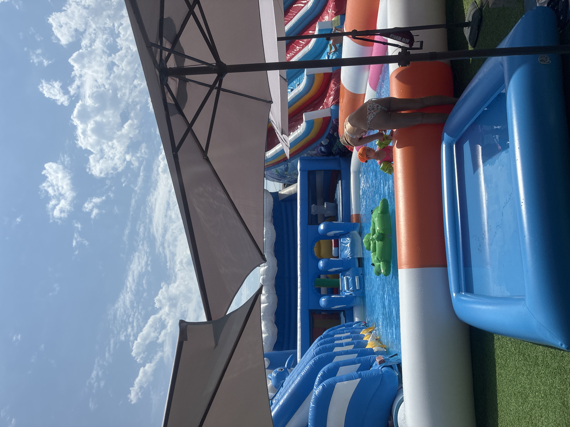 The image captures a vibrant scene at an inflatable water park, set against a backdrop of a bright blue sky filled with fluffy white clouds, suggesting a sunny day. In the foreground, a large, light-colored umbrella provides shade, its metallic frame visible.
The main focus is the water park, an expansive setup of inflatable structures in various bright colors. Dominating the right side is a large, blue inflatable pool or slide. To its left, a row of orange and white inflatable elements runs horizontally. Further back, a multi-colored inflatable slide, featuring red, white, blue, and rainbow sections, stands prominently.
Two people are visible in the water. On the right, a woman in a patterned bikini leans into the water, her legs extended. Near her, a young child wearing an orange swim cap and floaties is playing in the water. A green inflatable alligator floats nearby. Other parts of the water park include numerous blue and white inflatable structures, some with what appear to be climbing walls or slide entrances. Yellow buoys are visible floating on the water.
The overall impression is one of fun and recreation, likely during a summer day. The presence of the umbrella and the clear sky suggest it's daytime, probably mid-morning or afternoon. The artificial turf visible on the right side indicates a specially constructed recreational area. There is no visible text in the image, apart from what might be a small label on one of the orange inflatable elements that is partially obscured. The setting appears to be an outdoor amusement facility.