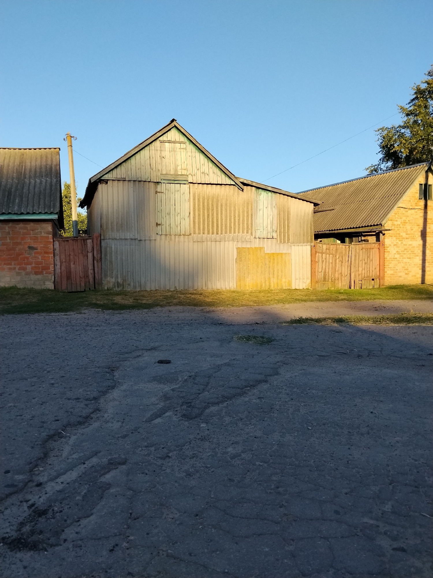 The image shows a view of several structures on a sunny day in Ustyvytsia, Ukraine. In the foreground, a worn asphalt road with visible cracks stretches across the frame. Behind the road, a grassy area is bordered by a corrugated metal fence. To the left of the fence is a brick building with a grey corrugated roof. Adjacent to it is a large structure with a sloped grey corrugated roof and walls covered in vertical corrugated metal siding and weathered wooden planks. This structure has a gabled wooden facade at the top, with a visible wooden door. To the right of this main structure is a smaller wooden gate. Further to the right, another building is visible, constructed of yellow brick with a grey corrugated roof and a small window. A utility pole stands to the left of the main structure, and a large tree with green foliage is visible behind the structures on the right side of the image. The sky is clear and blue.
