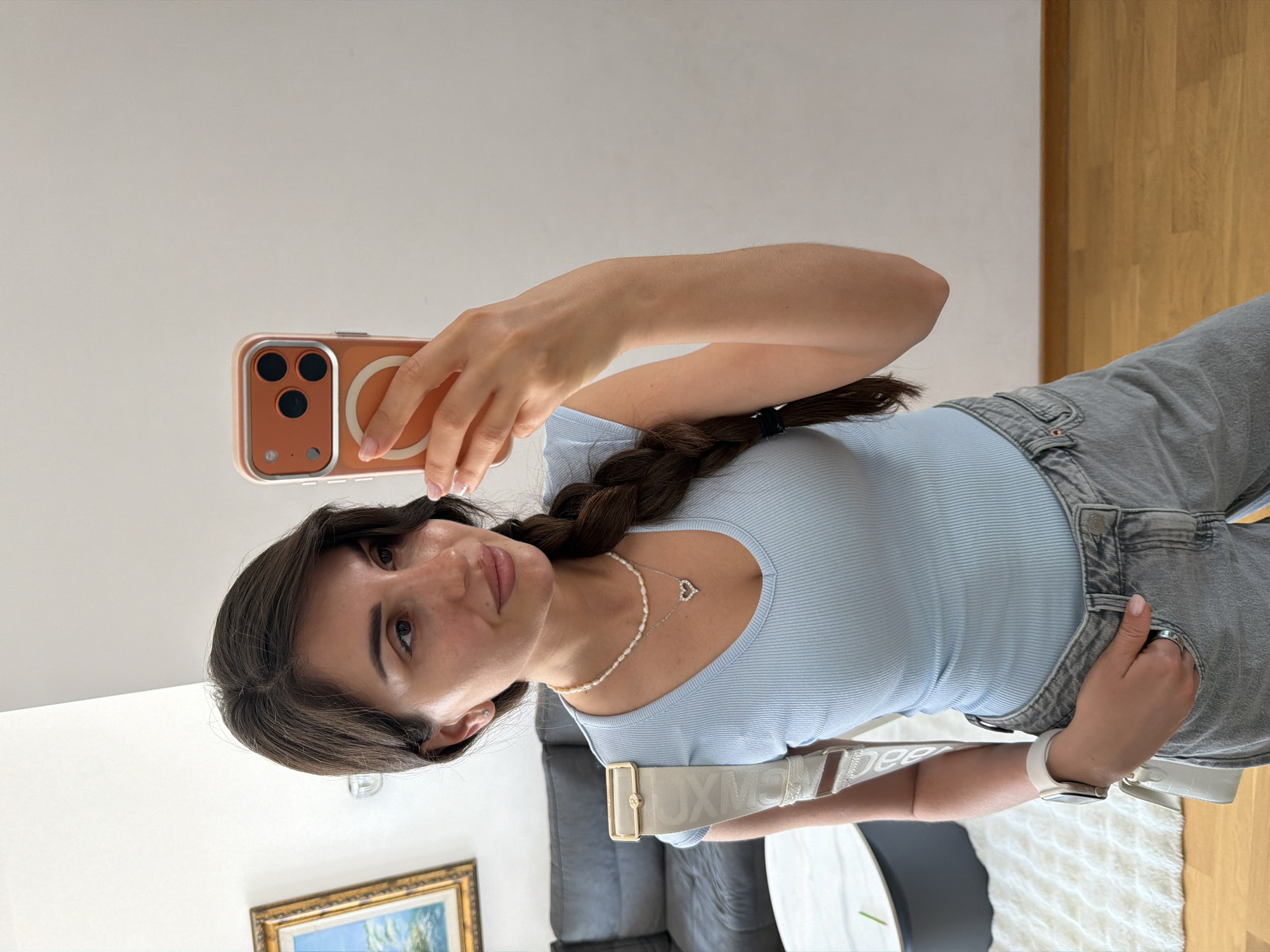 A woman is taking a selfie with a smartphone in a well-lit indoor environment. She has long, dark hair styled in a braid and is wearing a light blue fitted top, grey denim overalls, and jewelry consisting of a pearl necklace and a heart pendant. She is also wearing a watch on her left wrist. The phone she is using has a copper-colored case. A white purse is visible across her body.
Behind her, there's a light grey sofa, a round table with a dark surface, and a white shag rug. In the far background, a gold-framed painting is partially visible, positioned near the floor level. The floor is made of wood and appears to be well-maintained. The location is not explicitly identifiable as Budva, Montenegro.