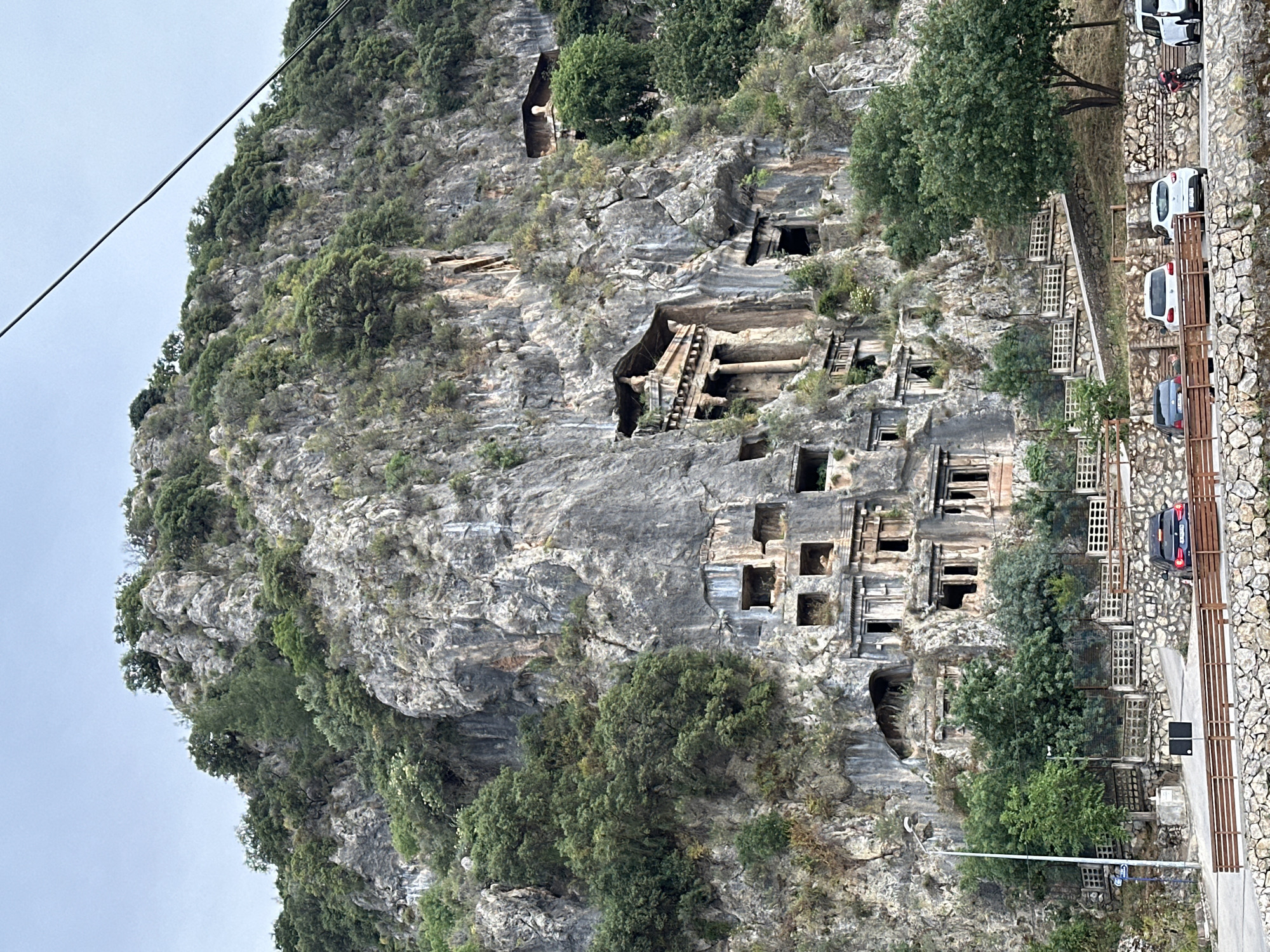 The image depicts the Lycian rock tombs in Fethiye, Turkey. The tombs are carved into a tall, rocky cliff face, interspersed with trees and vegetation. The rock face is grey and textured with visible geological strata. The tombs are square or rectangular openings, some with more elaborate architectural features like columns and facades. A road runs along the base of the cliff, with parked cars visible along the roadside. The sky appears overcast.