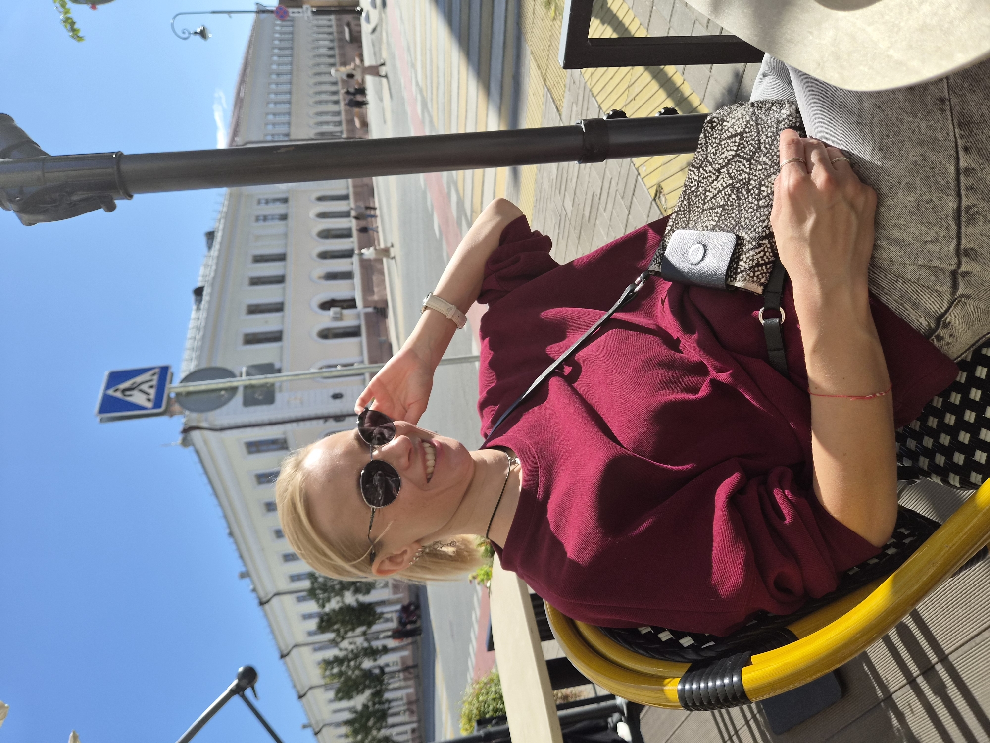 A woman with blonde hair pulled back and wearing sunglasses is seated at an outdoor cafe table on a sunny day. She is wearing a burgundy crewneck sweatshirt and light-wash jeans, with a patterned crossbody bag slung over her shoulder. She is smiling and touching her sunglasses. The cafe appears to be in Kazan, Russia, with a building in the background featuring multiple arched windows and an ornamental facade. A pedestrian crossing is visible on the street in front of the building. The woman is sitting in a yellow and black woven chair.