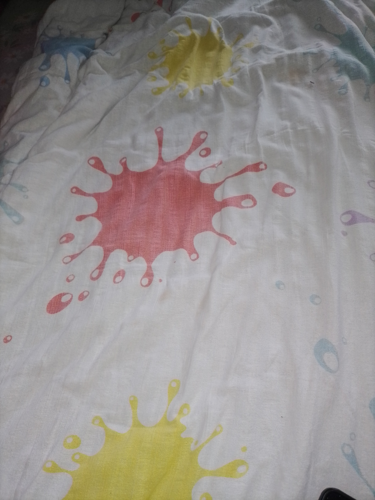 The image displays a white comforter with a pattern of colorful paint splatters. The splatters are in shades of pink, yellow, light blue, and purple, resembling splashes of paint. The comforter is creased and rumpled, suggesting it is either being used or has been stored in a disorganized manner. The lighting appears to be dim, possibly indicating an indoor setting with artificial light or a dimly lit room. The focus is on the textile surface and its abstract design. There are no people, specific objects, or discernible activities taking place. The context of Kalisz, Poland, does not provide any direct visual cues within the image itself.
