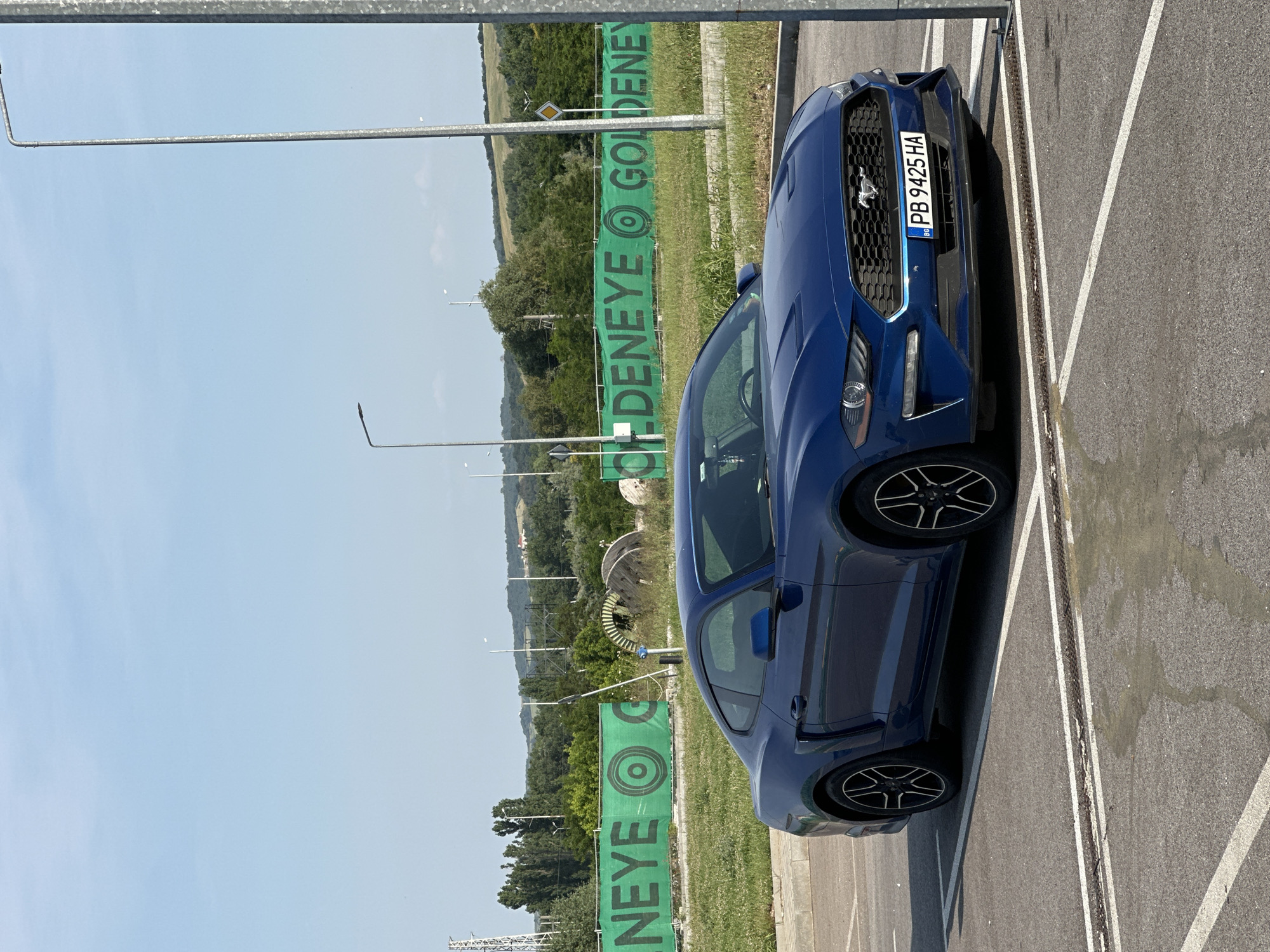 A dark blue Ford Mustang coupe is parked in an asphalt lot under bright daylight conditions in Kapitan Andreevo, Bulgaria. The vehicle is positioned facing forward and slightly to the left, with its front end near a vertical metal pole on the right side of the frame. The Bulgarian license plate "PB 9425 HA" is prominently displayed on the front bumper. The car features black multi-spoke alloy wheels. White painted lines delineate a parking space in the asphalt foreground, which also exhibits a wet or stained patch. In the midground, a long green mesh banner stretches horizontally, bearing the word "GOLDENEYE" repeated in black capital letters, interspersed with a circular target-like logo. Behind the banner, a grassy area contains various trees, shrubs, and large industrial-style wooden spools. A yellow rhombus-shaped road sign (priority road) is visible above the right side of the banner. Several tall, slender streetlights are present in the background, along with a distant power pylon on the far left. Rolling green hills form the horizon under a clear, light blue sky with minimal cloud cover.