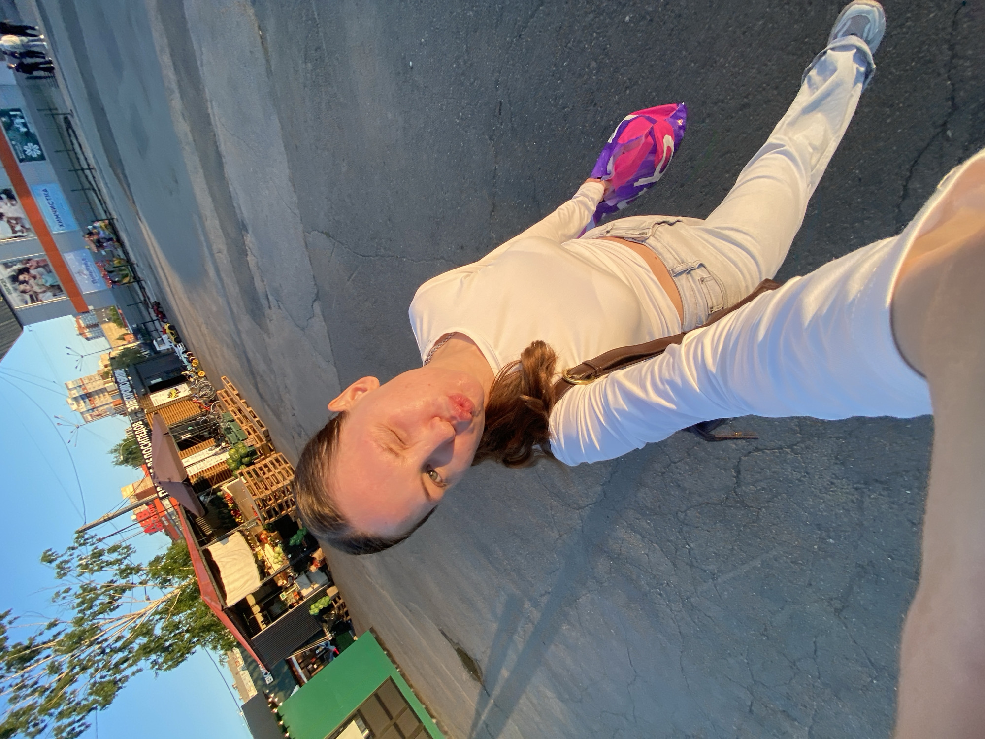 A selfie-style, low-angle, full-body shot depicts a young adult woman standing outdoors in a paved urban environment during what appears to be late afternoon or early morning sunlight. The woman, with brown hair pulled back, is making a winking expression with her left eye and pouting her lips. She is wearing a white long-sleeve crop top, light blue jeans, and light-colored sneakers. A brown shoulder bag is visible on her right side. In her left hand, she holds a vibrant pink and purple shopping bag with white Cyrillic text, possibly displaying "ТАКО" or a similar branding.
The background on the left features several market stalls or kiosks constructed from corrugated metal and wood, with produce such as fruits and vegetables visible on display and stacked on wooden pallets. A prominent green-sided stall is in the foreground. Deciduous trees with green foliage stand behind these stalls, and residential buildings are visible further in the distance under a clear blue sky.
The background on the right is dominated by a large, modern commercial building, possibly a shopping center, characterized by orange and grey facade elements. Prominent Russian signage is visible on its exterior: "ХИМЧИСТКА" (Khimchistka - Dry Cleaning) is clearly displayed. To its right, another sign partly reads "ВЕЛОСИПЕДОВ ЭЛЕКТРОМОПЕДОВ" (Velosipedov Electromopedov - Bicycles, Electromopeds), indicating a bicycle and electric moped sales or rental shop. Below the dry cleaning sign, an advertisement featuring children is visible. On the paved area in front of this building, a small, colorful children's amusement ride resembling a train and several miniature electric cars for children are present. A small group of people is standing near the building. The ground is asphalt with visible cracks.
Based on the Russian signage and architectural style, the scene is located in Izhevsk, Russia, likely near the Avangard Shopping Center on Pushkinskaya Street, an area known to feature both informal market stalls and a commercial complex with various shops and services.