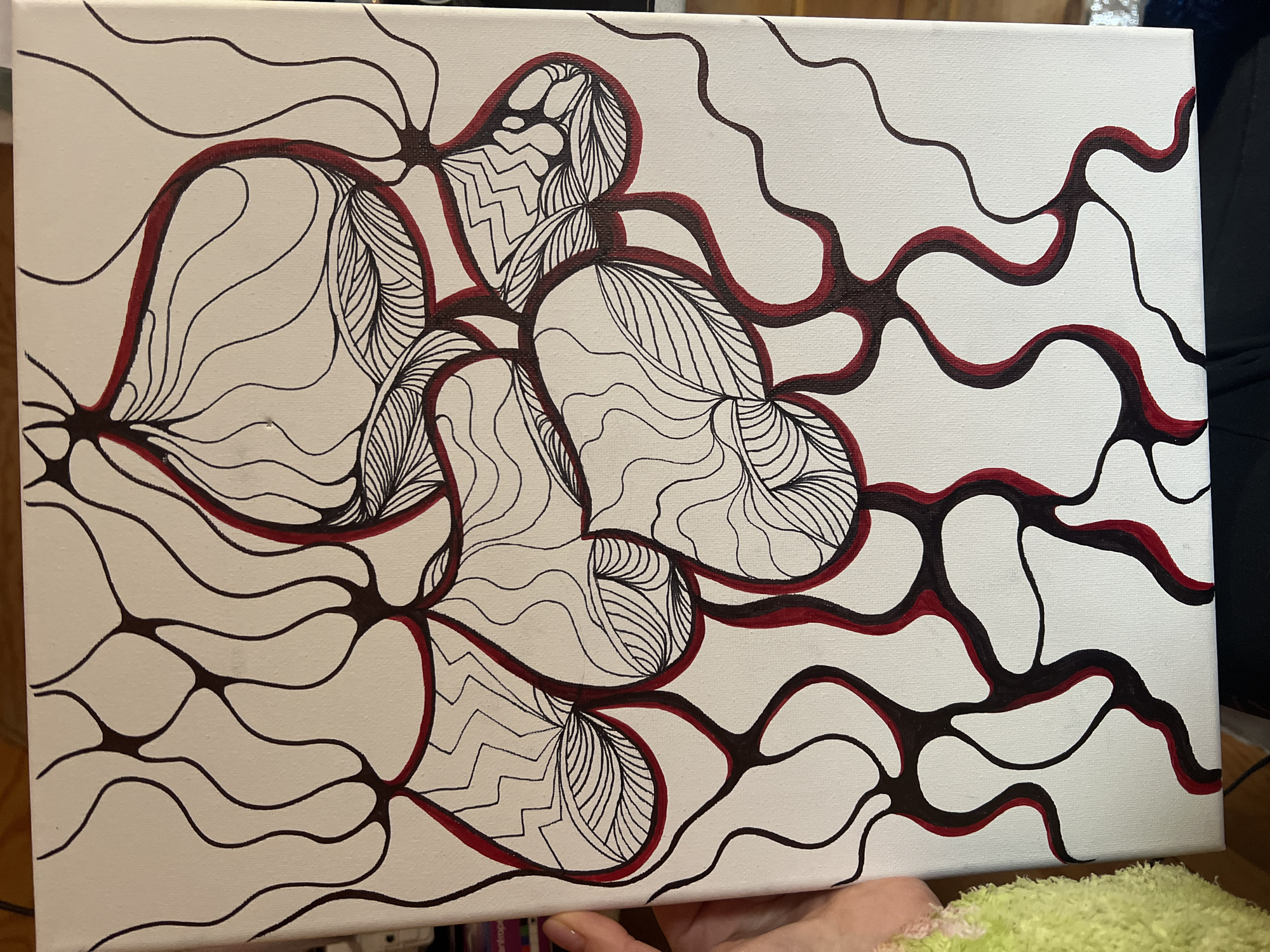 An abstract painting on a white canvas, held by a person's hand, is the central subject of the image. The artwork features an intricate network of flowing, organic lines primarily in black, with some lines and shapes outlined or filled with a dark red hue. These lines interconnect, forming bulbous nodes and larger, heart-shaped or leaf-like forms.
Within these larger shapes, various intricate patterns are drawn in black ink, including wavy lines, parallel stripes, and zig-zag motifs, creating a doodle-like or Zentangle aesthetic. The canvas texture is visible, indicating a painted or drawn medium rather than a digital print.
The activity depicted is the presentation or photographing of this artwork. A human hand is visible in the lower right corner, holding the canvas at a slight angle. Part of a fluffy, light green garment or blanket is also visible at the very bottom edge of the frame. The background behind the painting is out of focus and appears dark, suggesting an indoor setting. There is no visible text on the artwork or in the immediate surroundings. The lighting is even, highlighting the details and colors of the painting.