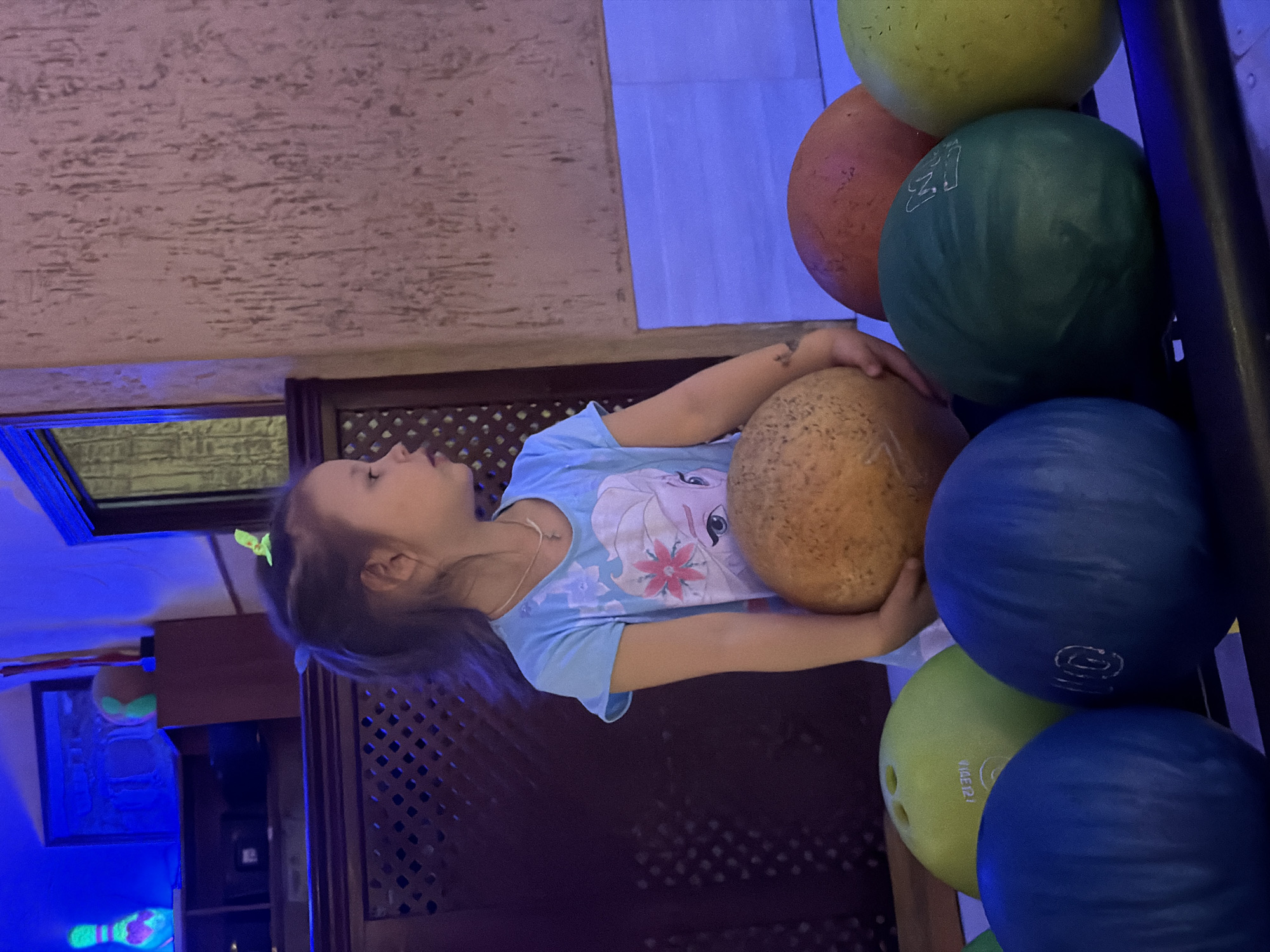 A young girl is holding a bowling ball in what appears to be a bowling alley. She is wearing a light blue t-shirt with an image from the movie "Frozen" on it. Her hair is tied back with a light green hair tie. She is surrounded by a bowling ball rack containing bowling balls in various colors including yellow, orange, green, and blue. The background shows a textured wall and what looks like a decorative, dark wooden structure behind her. There is blue lighting, and what appears to be a bar area or seating area to the left with lit up bowling pins and other items. It is located in the city of Chaikovsky, Russia.