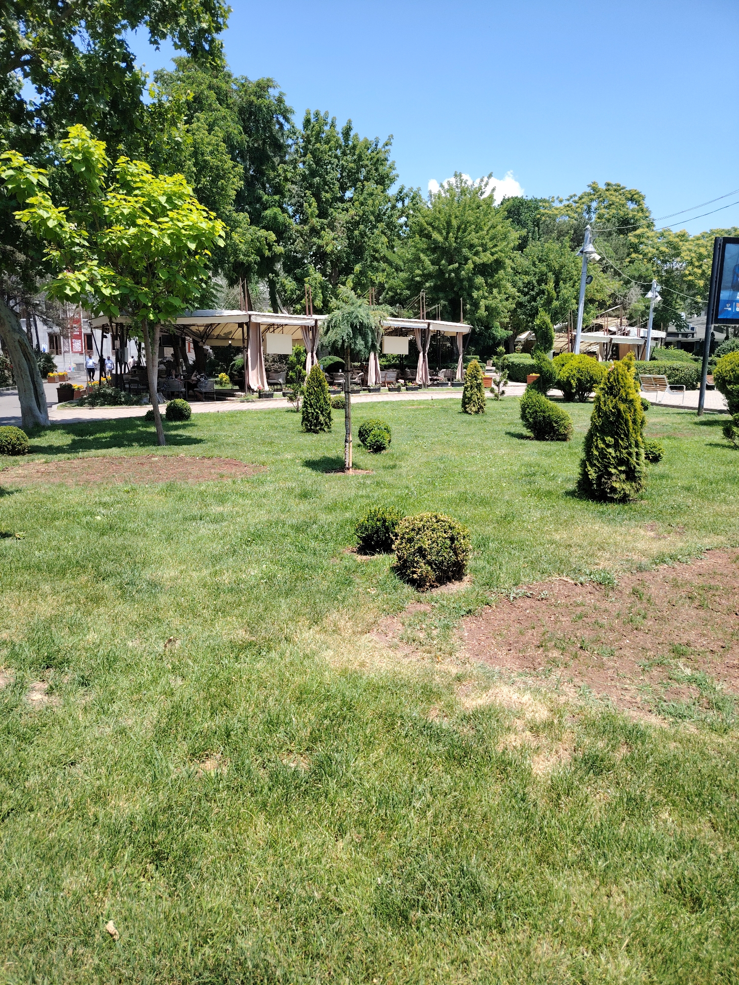 The image depicts a sunlit park or garden area with manicured green grass and various trees and shrubs. In the background, there are several outdoor dining gazebos with white canopies, suggesting a cafe or restaurant. A few people are visible walking on a path behind the gazebos. The sky is clear and blue. The presence of a large billboard on the right side of the image, which is common in urban environments, and the general landscaping suggest a public space. This scene is consistent with the Tumo Park in Yerevan, Armenia, known for its green spaces, outdoor seating areas, and artistic installations.