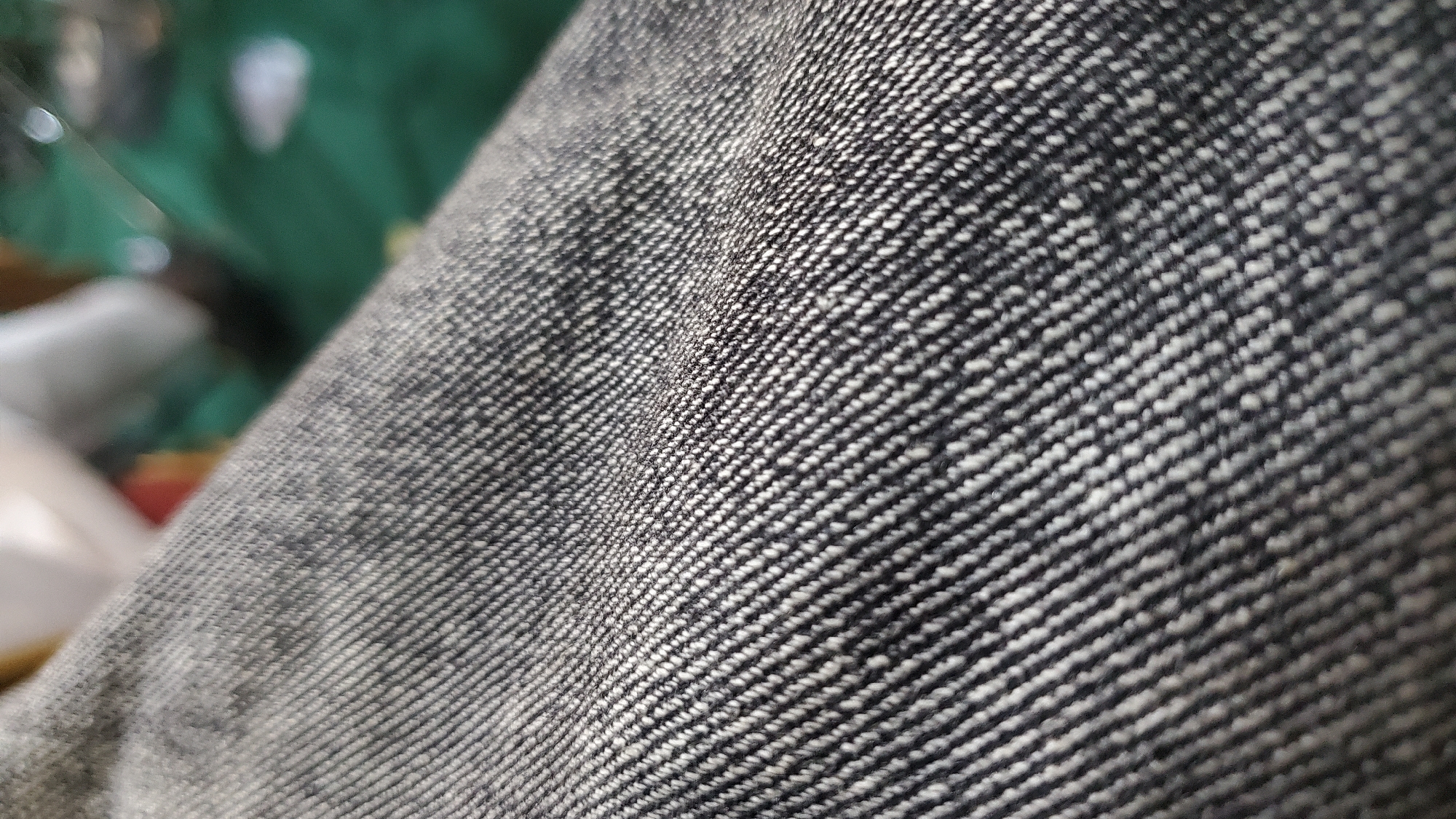 The image shows a close-up, angled view of dark denim fabric. The texture of the weave is clearly visible, with diagonal lines of dark and lighter threads creating a ribbed effect. The fabric appears to be slightly creased, suggesting it may be part of clothing or upholstery. The background is out of focus, but a blurry green surface and some lighter, indistinct shapes are visible, hinting at an indoor or possibly outdoor setting with some other objects present. There are no people or explicit signs of activity. There is no information available to determine the location within Jalingo, Nigeria.