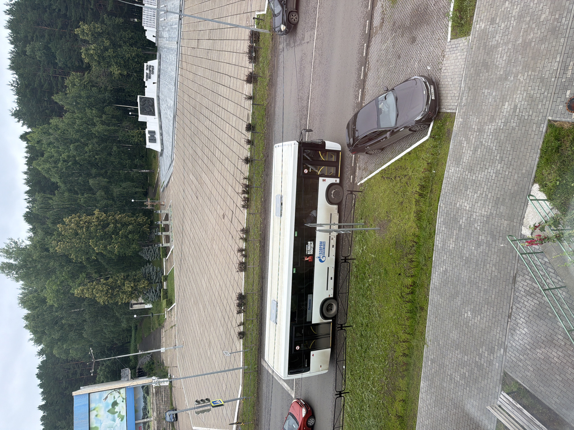 In the city of Chaikovsky, Russia, on a cloudy day, a white bus with "GAZPROM" and "VELIKAYA POBEDA!" (Great Victory!) text is stopped at a bus stop. Next to it, a red car is also stopped. A dark gray car is parked in a lot on the right. The area is paved with gray cobblestones and surrounded by green grass and trees. In the background, a memorial with white stone structures and trees can be seen. A traffic light is visible on the left.