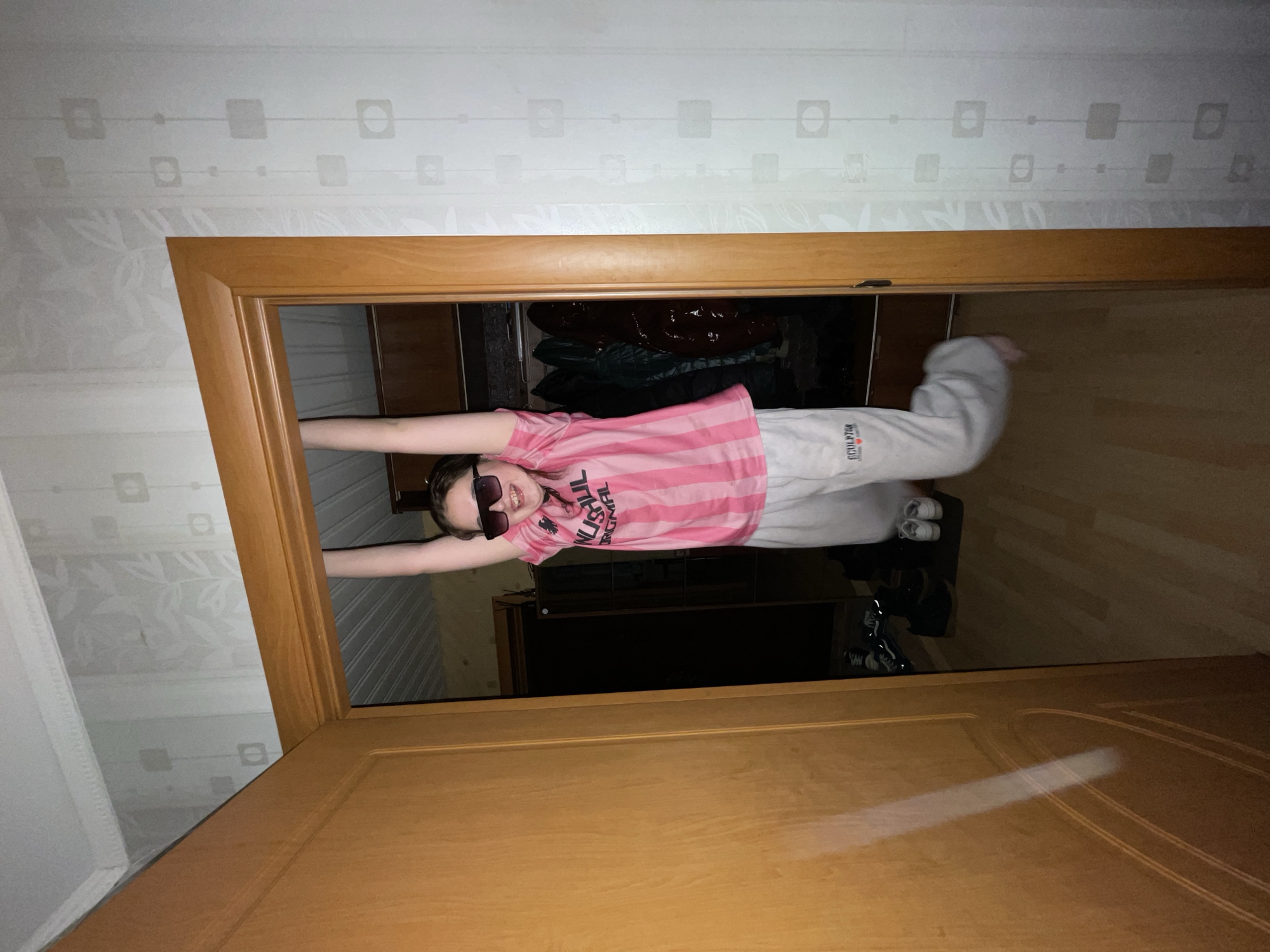 An indoor photograph, originally rotated 90 degrees, depicts a young person playfully hanging horizontally within a wooden door frame. The individual, likely a girl or young woman, is centered in the doorway, gripping the top of the frame with outstretched arms, suspending her body in mid-air. She is wearing a pink and white striped t-shirt with the partial text "UNUSUAL ORIGINAL" visible on it, light grey sweatpants with "SCULPTOR" visible near the left ankle, and dark sunglasses. She is smiling broadly at the camera, suggesting a cheerful and acrobatic pose.
The setting is an interior space in Murino, Russia. The door frame is made of light-colored wood. Around the doorway, the walls are covered in light wallpaper with a subtle pattern featuring squares, circles, and faint floral designs. A partially visible, light wooden door is in the foreground, showing a bright reflection of light. Through the open doorway, another room is visible, featuring a dark floor (possibly wood or laminate) and dark furniture, which appears to be a wardrobe or cabinet. A pair of dark sneakers with white soles are on the floor in this background room. The lighting is artificial and somewhat dim, creating reflections on the wooden surfaces. The overall mood is lighthearted and playful.
