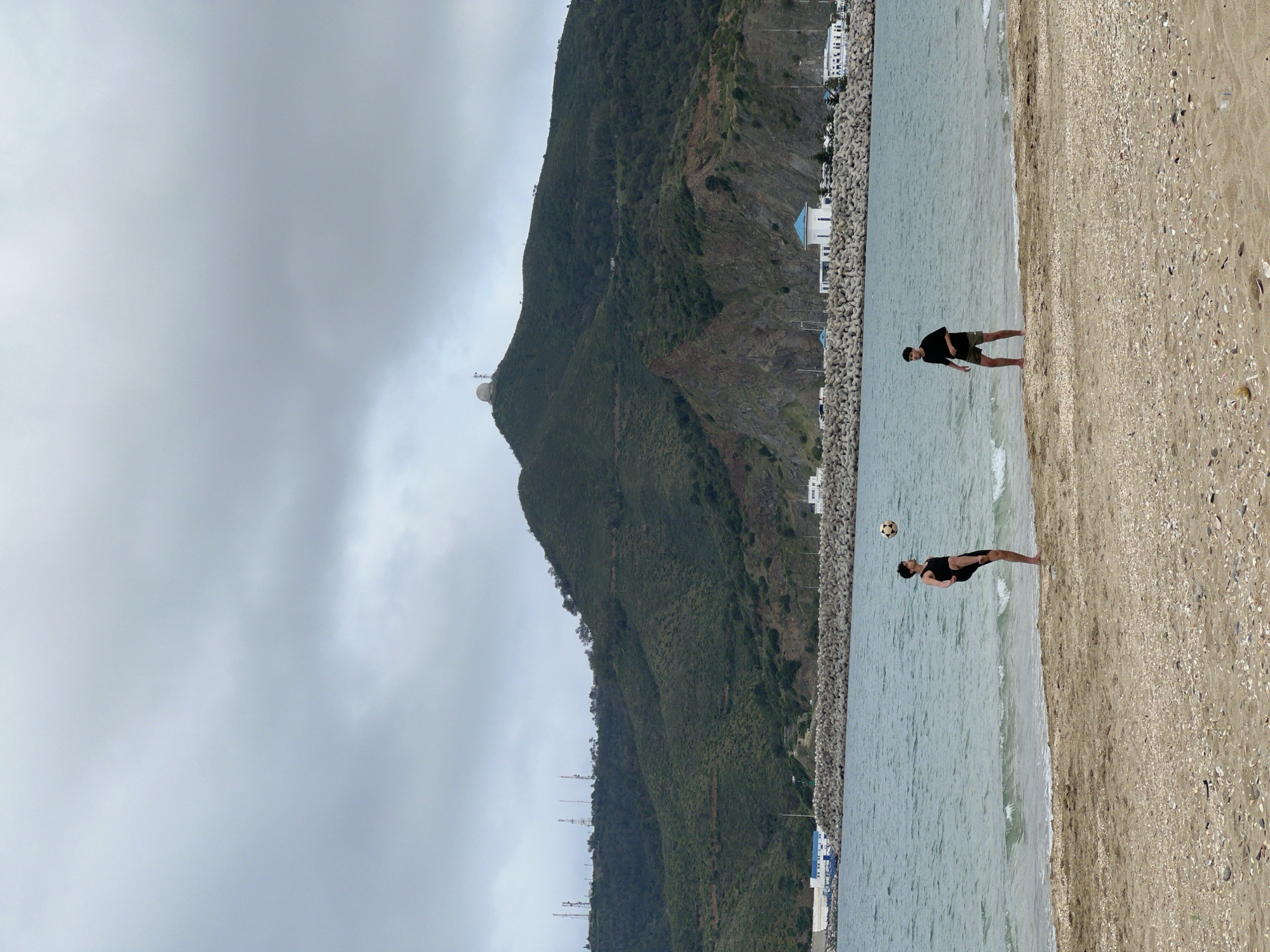 Two people are playing soccer on a beach in M'Diq, Morocco. A mountain looms in the background, with a white dome visible at its peak. The sky is overcast. The beach has a mix of sand and shells. A sea wall made of concrete blocks runs parallel to the shore. Buildings can be seen beyond the sea wall. One person is kicking a soccer ball mid-air. Both are on the shoreline where the water meets the beach.