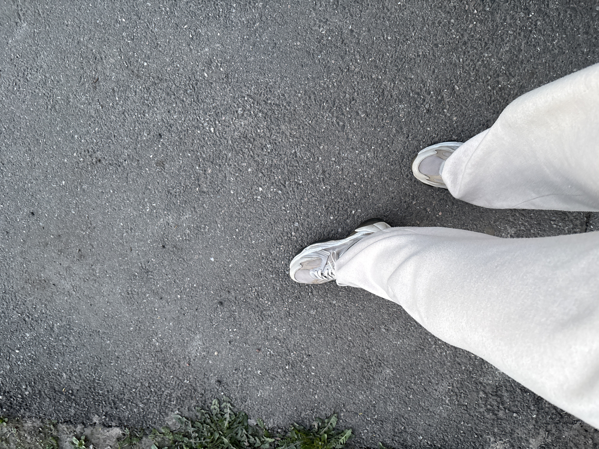 The image shows a first-person view of a person standing on asphalt. The lower portion of the person's legs is visible, clad in light-colored, possibly white or light gray sweatpants and gray sneakers. The asphalt surface is typical of city streets, showing a mix of fine and coarse aggregate. Along the lower left edge, there is vegetation, suggesting a roadside or edge of a pavement area. The specific location is not visually determinable as Saint Petersburg, Russia, without additional contextual information in the image.