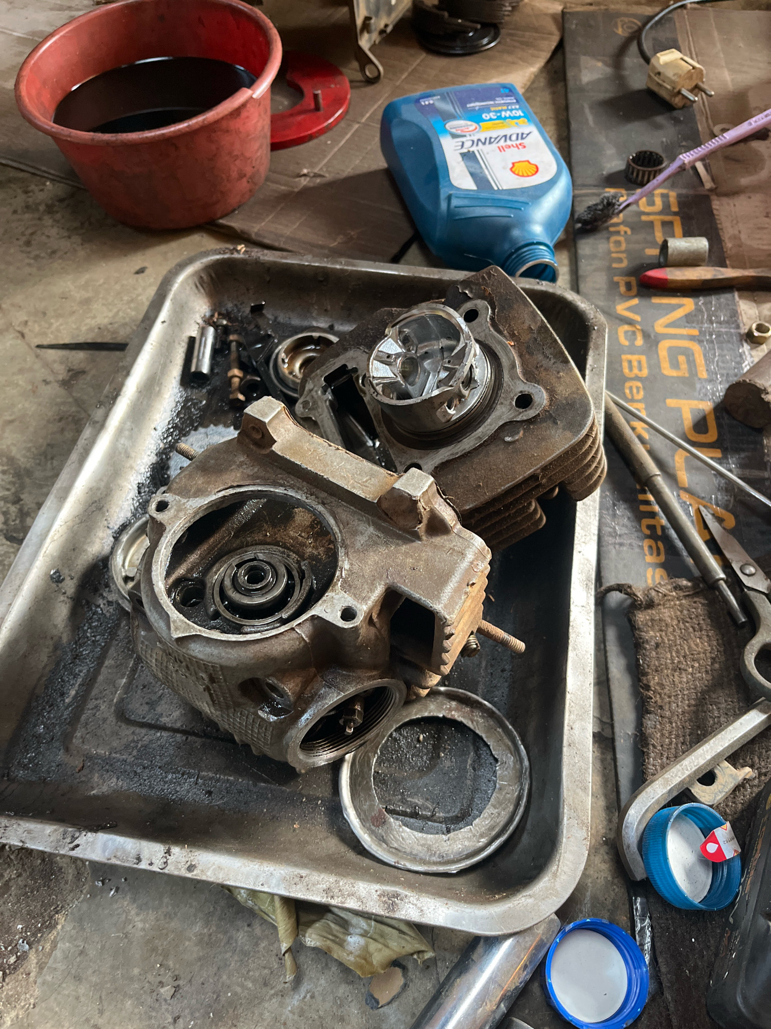 An overhead shot depicts a close-up of motorcycle engine parts, specifically cylinder heads, resting in a metal tray. The parts are dirty and appear to have been recently disassembled. Several tools and automotive fluids are scattered around the tray. In the background, a red plastic container holds dark liquid, likely used engine oil. A blue bottle of Shell Advance motorcycle oil is also visible, along with various small metal components, scissors, and a wrench. The overall scene suggests a mechanic's workspace, possibly in an outdoor or open-air setting given the rough concrete floor. The image does not provide specific visual cues to pinpoint its location within Kebumen, Indonesia, other than general context of mechanical work.