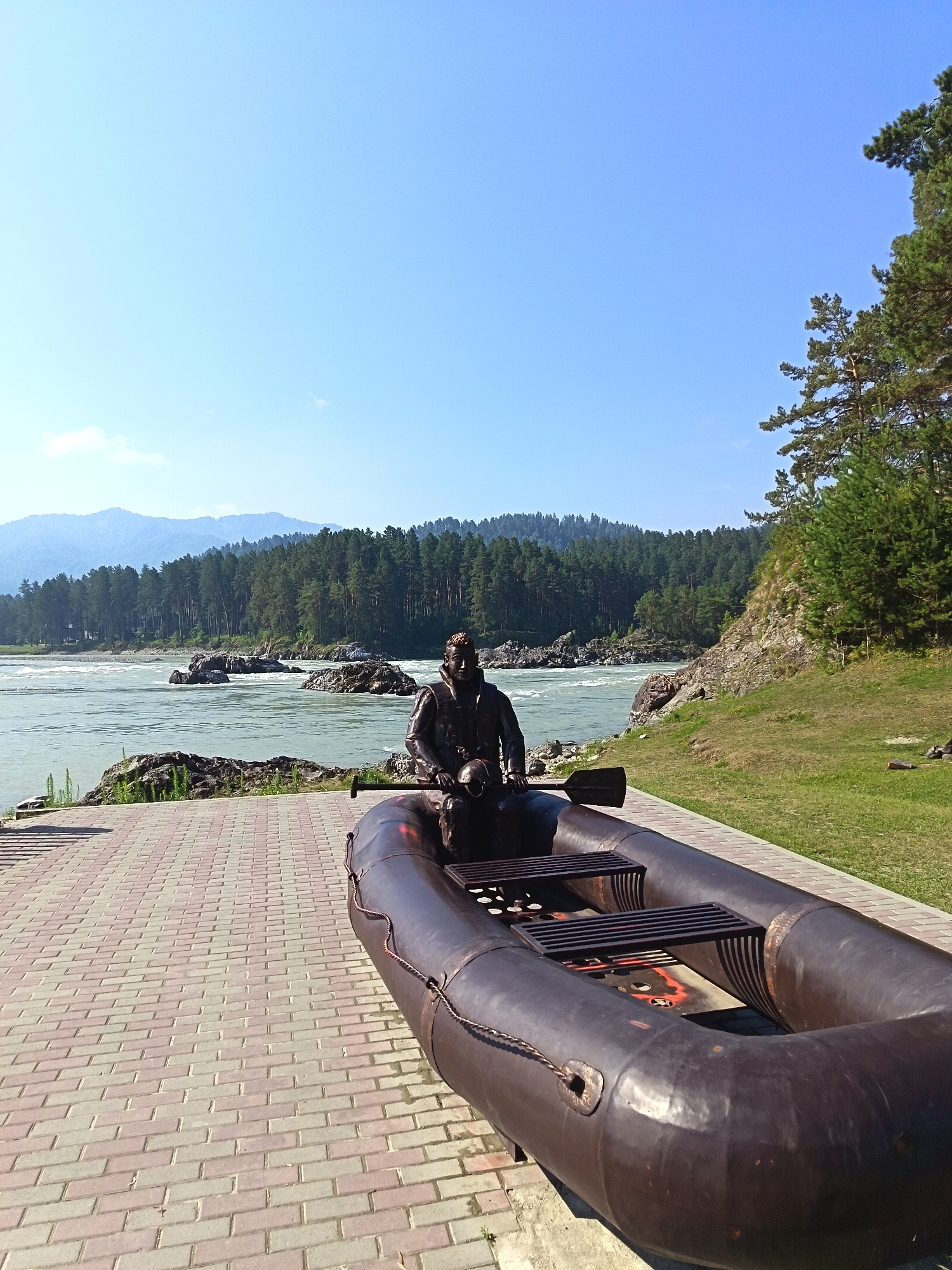 The image depicts a bronze statue of a man in a life vest sitting in an inflatable raft. The raft is positioned on a paved walkway made of interlocking bricks. Behind the raft, a river flows with visible rapids and large rocks in the water. The river is bordered by a dense forest of evergreen trees, and in the distance, mountains are visible under a clear blue sky. The scene appears to be a park or recreational area, likely located in or near Manzherok, Russia, given the natural landscape. The statue and raft are the primary subjects in the foreground, with the river and forest forming the background. The lighting suggests a bright, sunny day.
