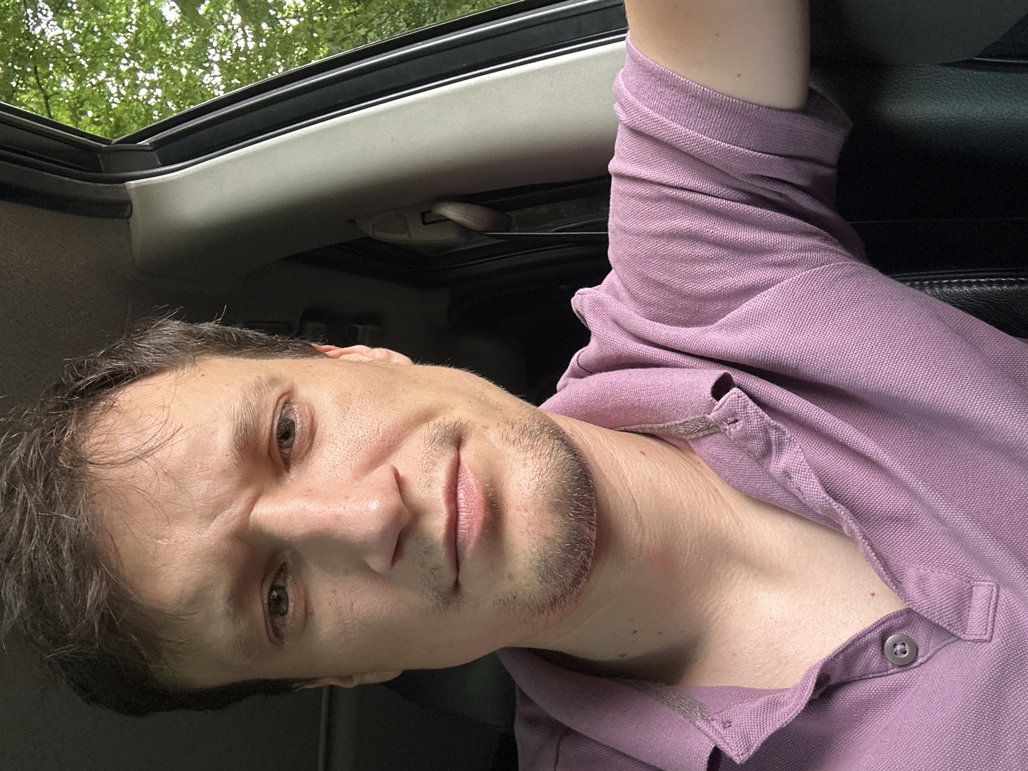 The image is a close-up, selfie-style photograph of a man in a vehicle, likely taken from the driver's seat. The man has short, dark hair, brown eyes, and appears to be in his late 20s to early 40s. He has a slight stubble on his chin and a faint smile. He is wearing a purple polo shirt with a collar. The interior of the vehicle is visible, including a black leather seat and a portion of the door frame and window. Outside the window, green trees and foliage suggest an outdoor, possibly wooded, environment. The image does not contain any information that could be used to determine the specific location in Shatava, Ukraine.
