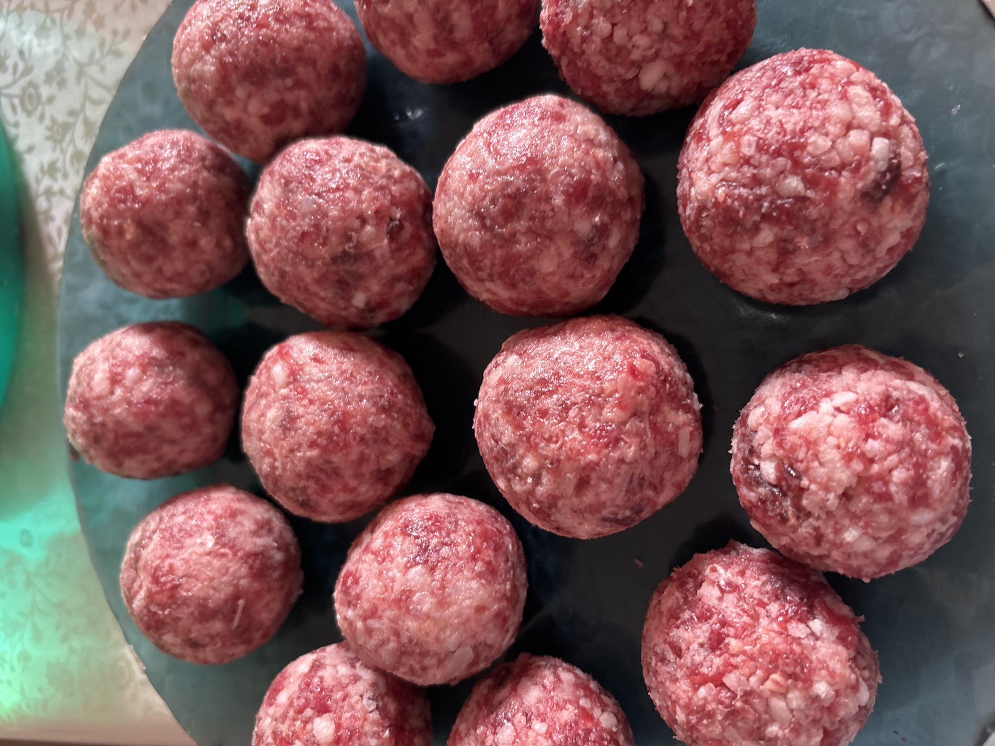 The image displays a close-up, overhead view of approximately 18 uncooked meatballs arranged on a dark teal plate. The meatballs are round and appear to be made of ground meat, with visible flecks of fat and possibly other ingredients. The lighting suggests an indoor setting, with a hint of a patterned tablecloth visible in the blurred background. There are no people, actions, or interactions depicted in the image. The scene is static and focuses solely on the raw food preparation. There is no information in the image that can be used to determine its location in Churapcha, Russia.