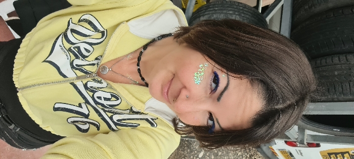 A woman is depicted in a close-up, mid-shot photograph, gazing directly into the camera with a slight smile. She has dark brown, chin-length hair with a center part. Her left eye features blue winged eyeliner, and iridescent green glitter is applied to her left cheekbone. She has a silver stud piercing on her left nostril and a silver hoop through her septum. She is wearing a yellow, cropped, zip-up hooded sweatshirt with the words "Detroit '76" printed in dark letters on the chest. A partially unzipped silver zipper is visible. Around her neck, she wears a black beaded necklace with silver accents, and below it, two silver necklaces, one with a circular tree-of-life pendant. Fine silver glitter is visible on her upper chest. In the background, out of focus, stacked black vehicle tires are visible on a metal rack to her left. To her right, a portion of a white utility van with yellow markings, including "052" and a red taillight, is discernible. The ground beneath the tires appears to be gravel or crushed stone. This scene is situated in an outdoor area, likely a service garage or tire shop, within the Mate Asher Regional Council, Israel.