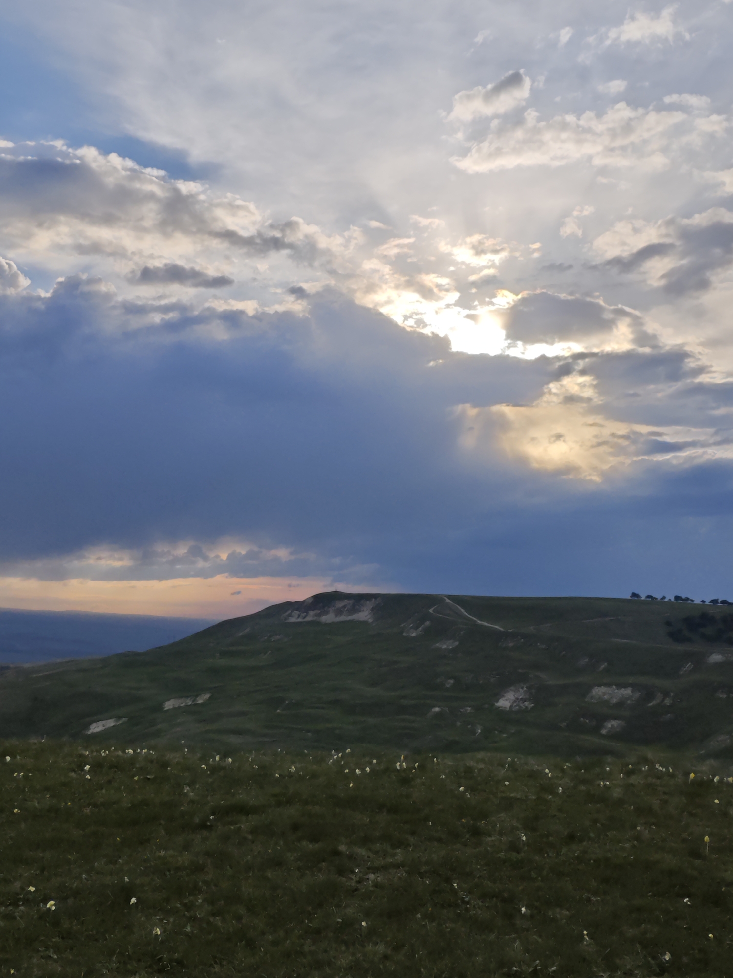 The image depicts a landscape with a grassy field in the foreground and a rolling hill in the middle ground. The sky is filled with clouds, some of which are dark and ominous, while others are lighter and illuminated by the sun. There is a hint of sunset or sunrise in the distance where the sky meets the horizon. The location and the exact city are unknown.
