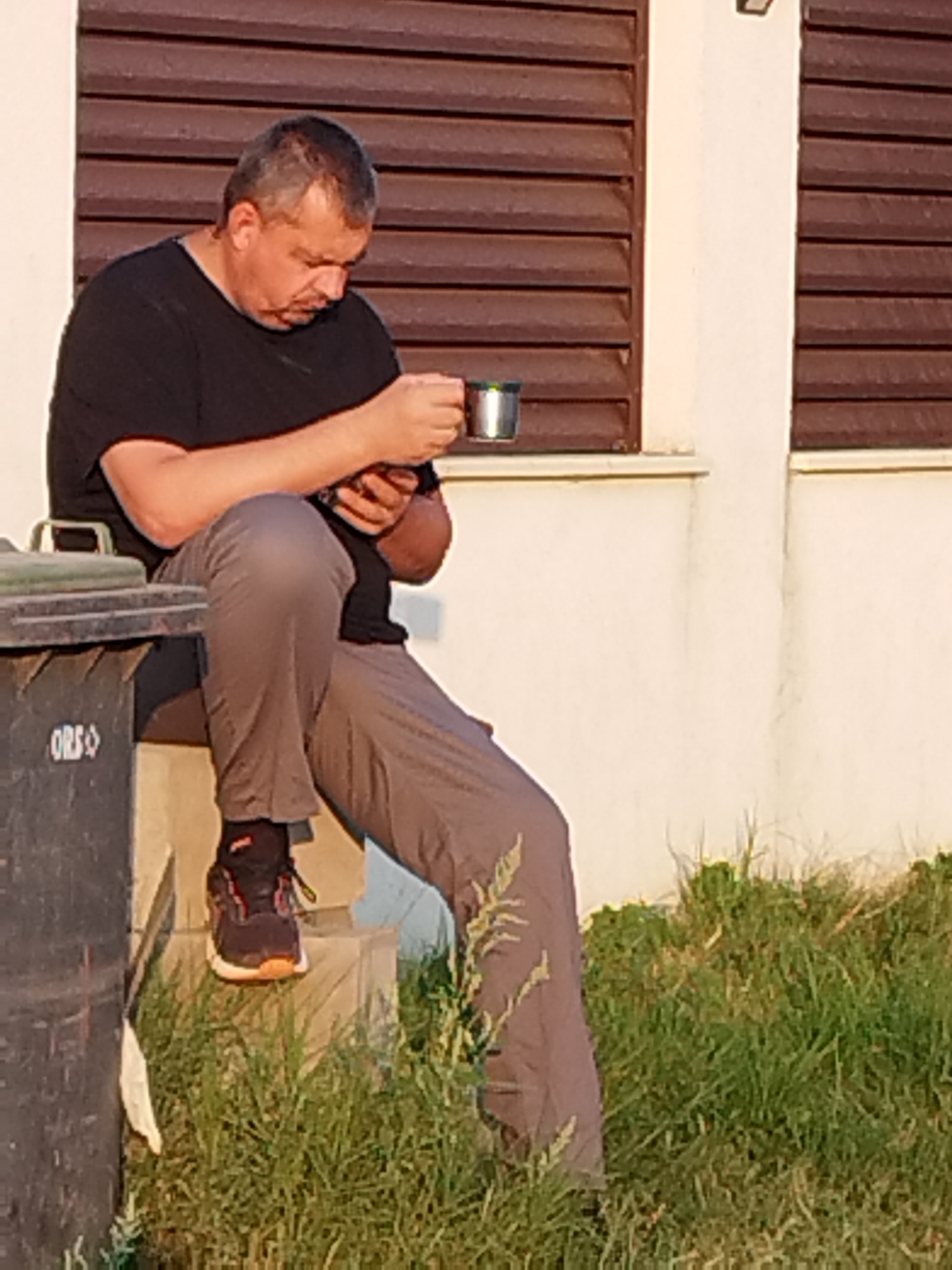 A middle-aged man with short, graying hair and a black t-shirt is sitting on a concrete step, holding a metal cup in his right hand and looking down at a mobile phone in his left. He is wearing gray pants and dark athletic shoes with orange accents. To his left, a large black garbage bin with the word "ORS" is visible. The background features a cream-colored wall with horizontal brown shutters on windows, and green grass in the foreground. The lighting suggests it is late afternoon or early evening. This scene is located in Vidra, Romania.