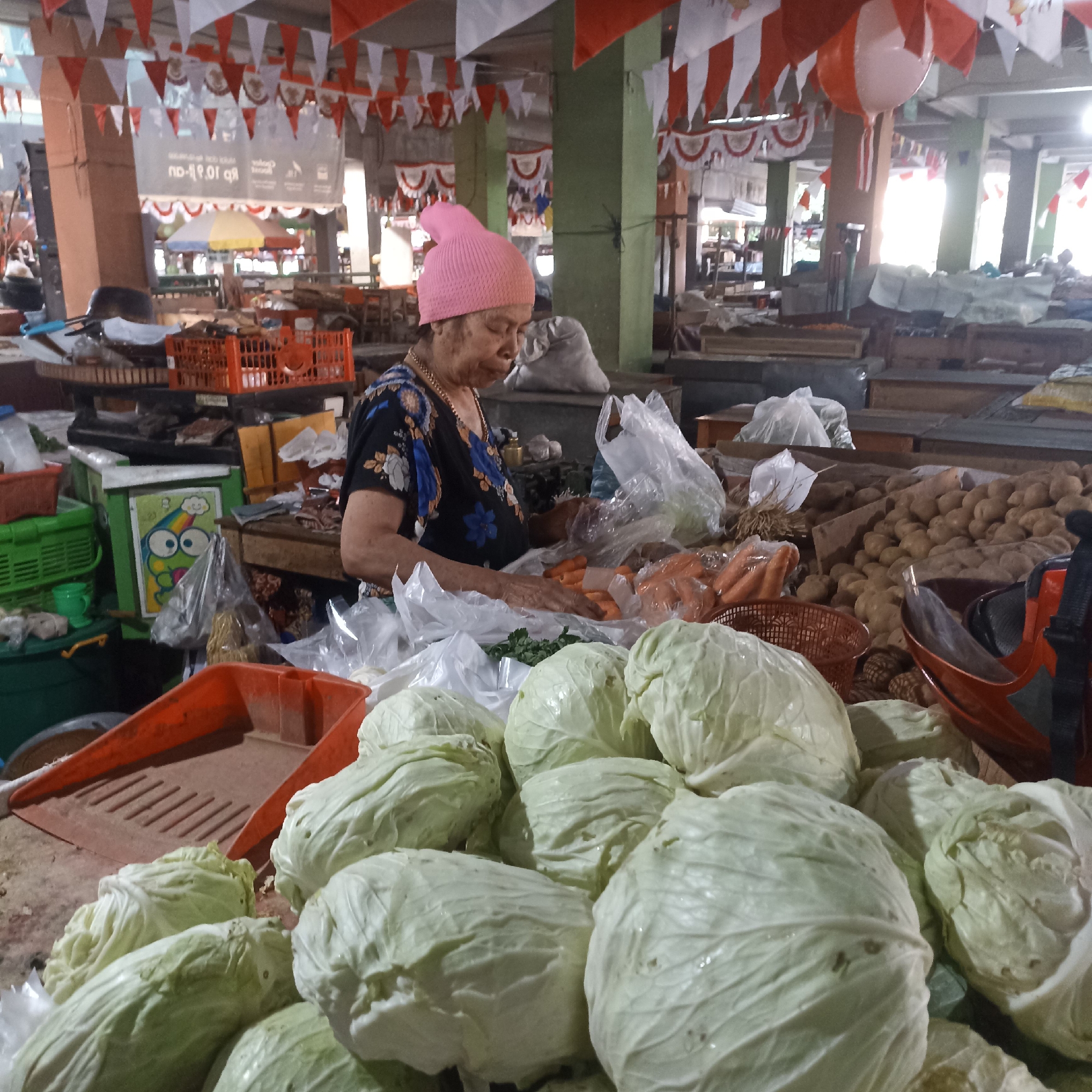 An elderly woman, wearing a pink knit cap and a floral print shirt, stands at a market stall in Magelang, Indonesia. She is in the process of preparing vegetables, her hands reaching towards a pile of chopped carrots and leafy greens. The stall is filled with produce, including large heads of cabbage prominently displayed in the foreground, along with carrots and potatoes. Various plastic bags and baskets are scattered around the stall, holding other items for sale. Overhead, red and white triangular pennants are strung across the market, indicating a festive occasion. The background reveals the bustling environment of the market, with other stalls and shoppers visible in the distance, under a covered roof supported by pillars.